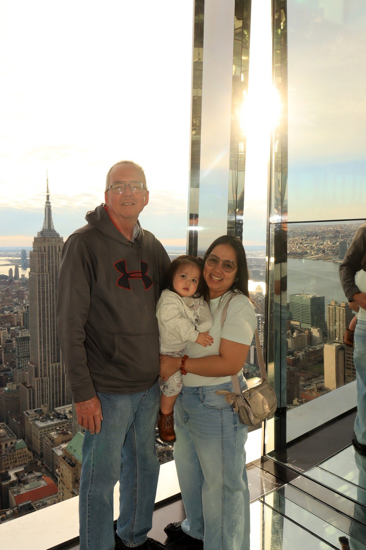 A family of three standing inside a skyscraper with a view of New York City, featuring the Empire State Building in the background, during sunset.