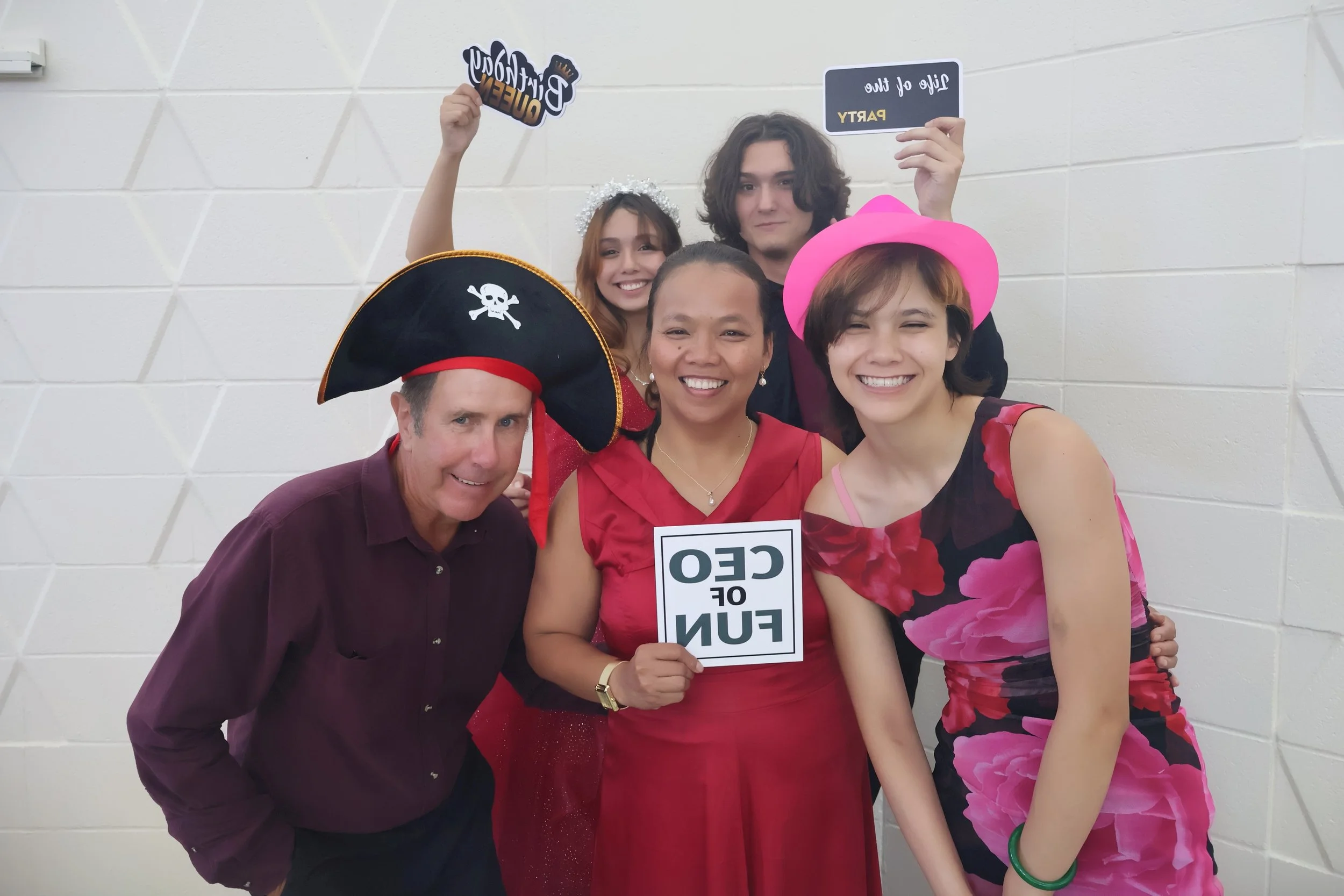A group of six people dressed festively standing close together against a white wall, some holding signs and wearing costumes, smiling at the camera.