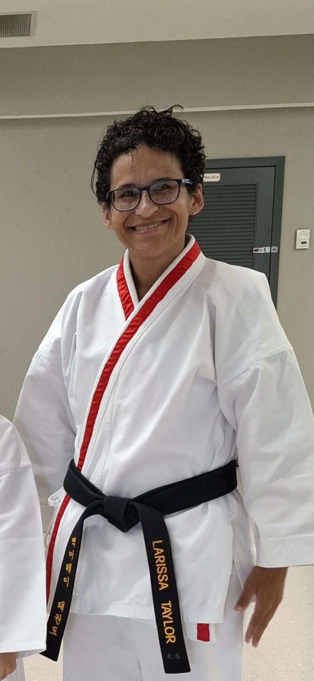 Person smiling in a karate uniform with a black belt, glasses, and curly dark hair.