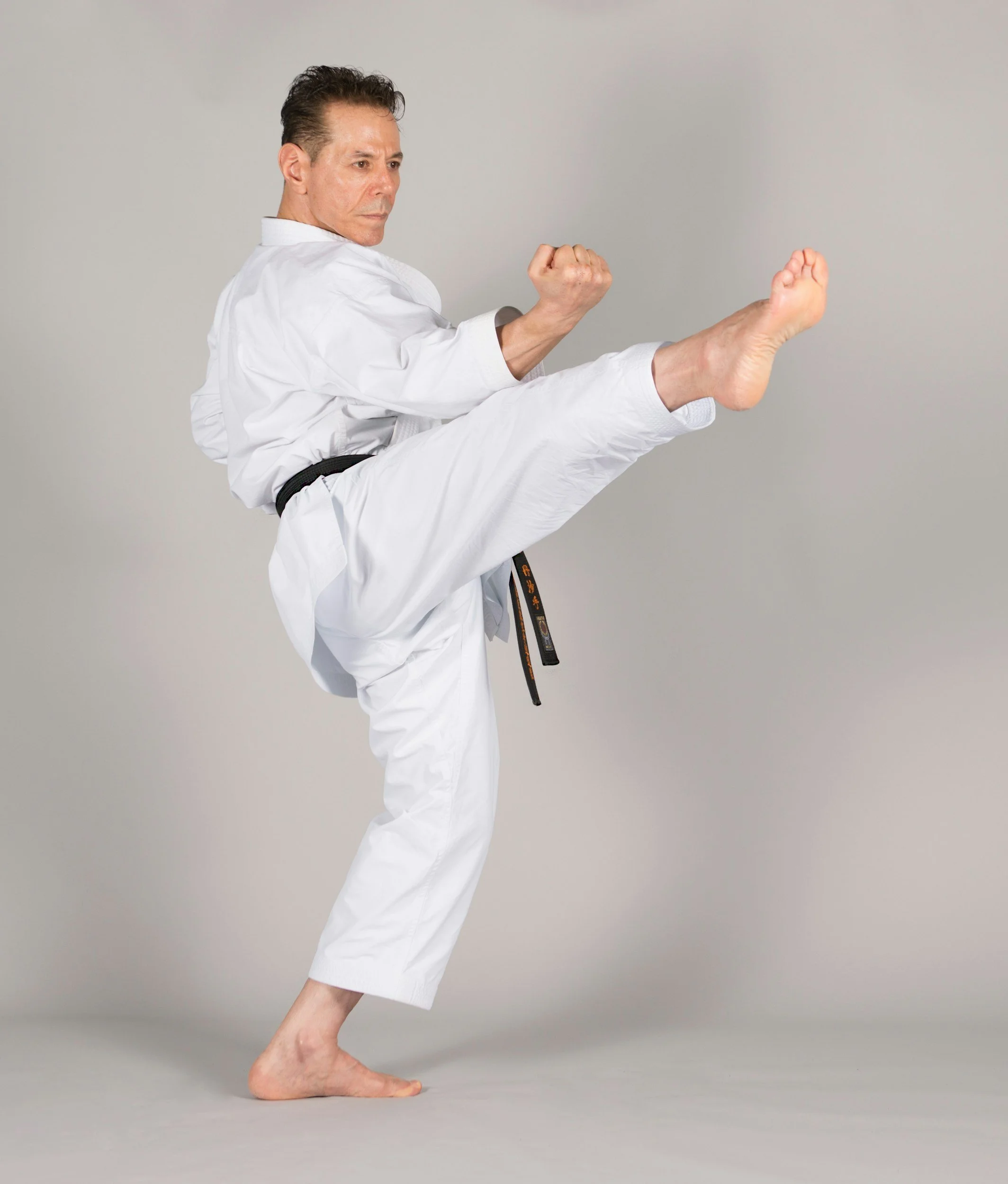 A man dressed in a white karate uniform performs a high front kick, balancing on one foot, against a plain gray background.