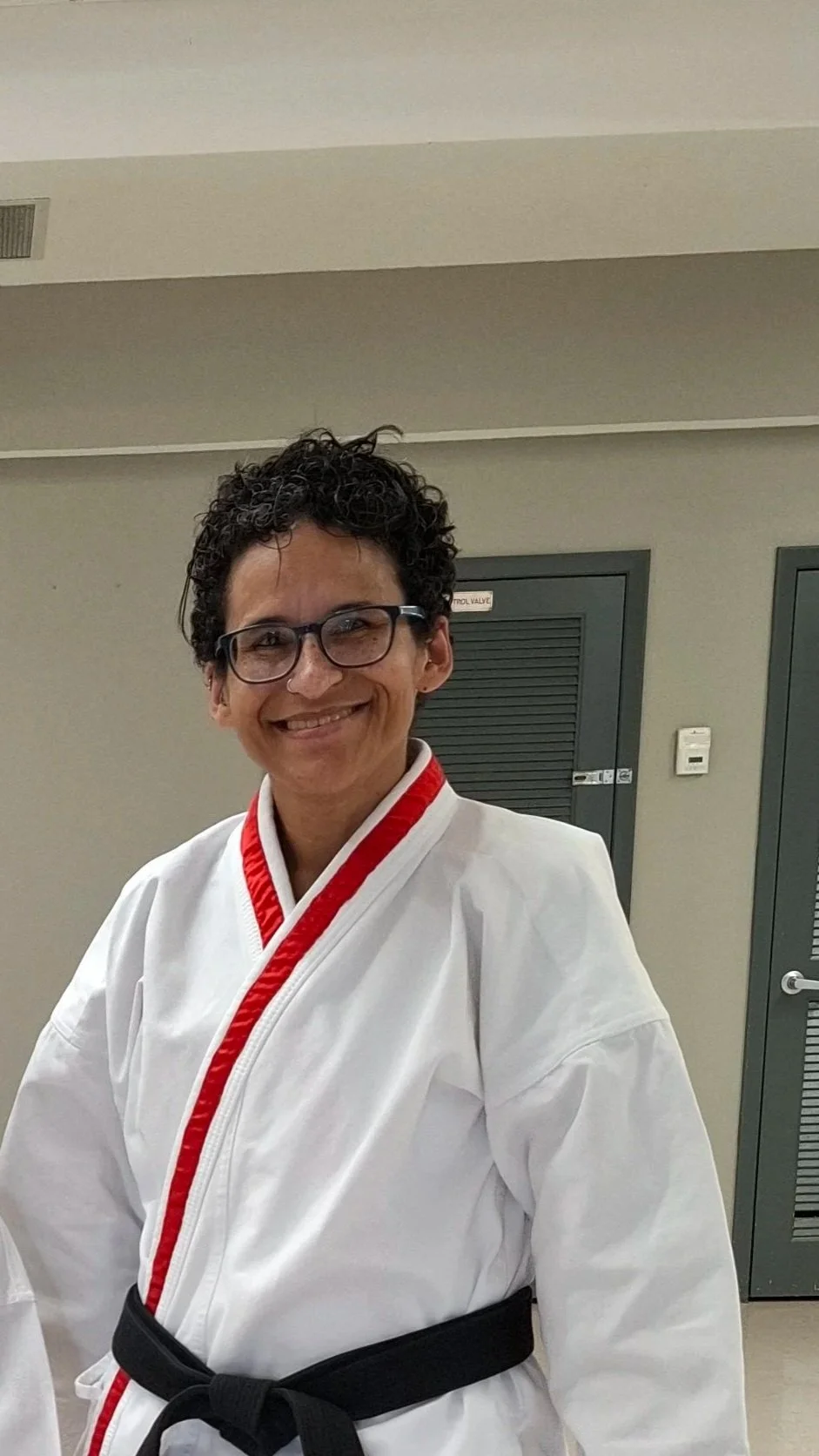 A woman with short curly hair and glasses, smiling, wearing a white martial arts gi with red trim and a black belt, standing indoors.
