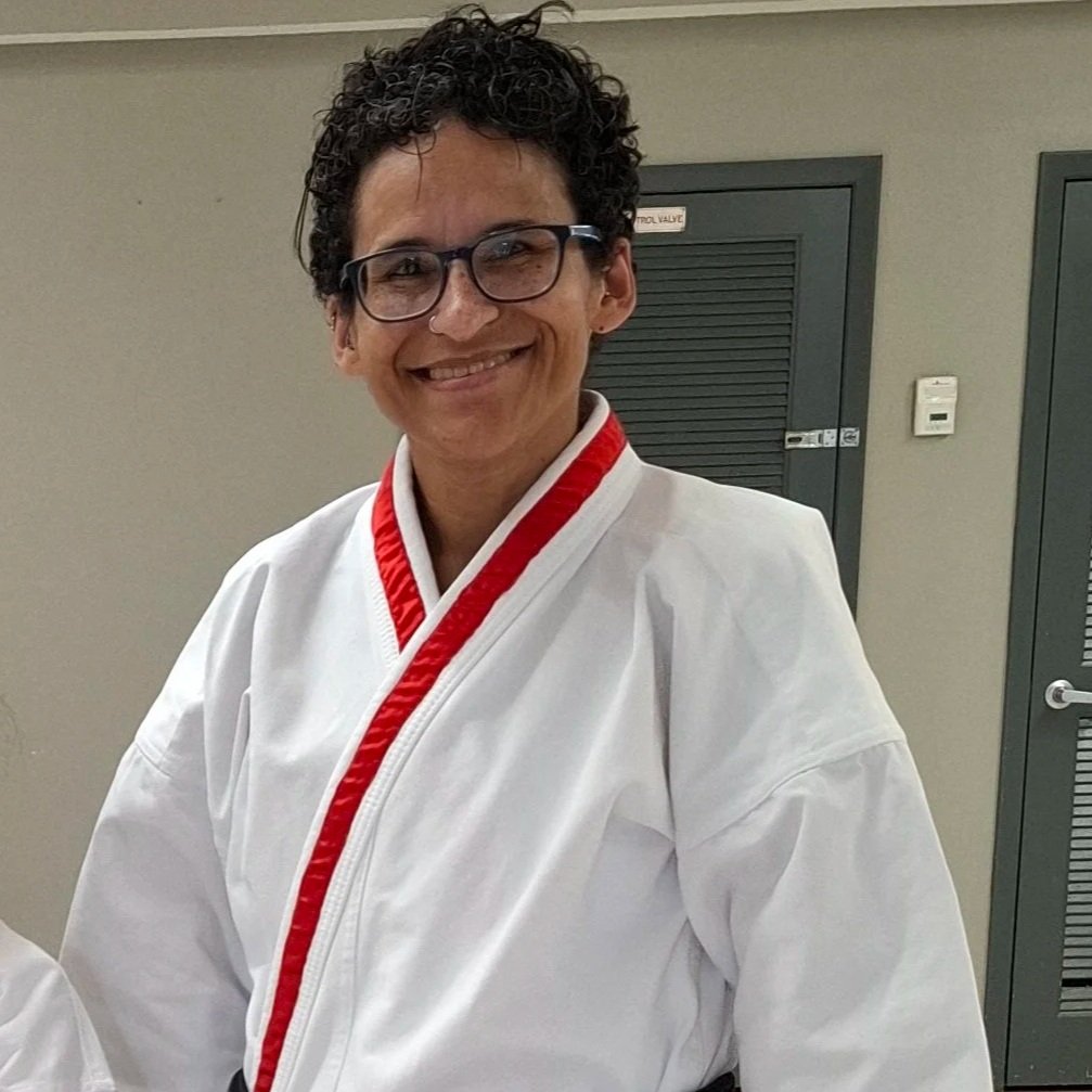 A person with curly dark hair, glasses, and a big smile wearing a white martial arts uniform with red trim.