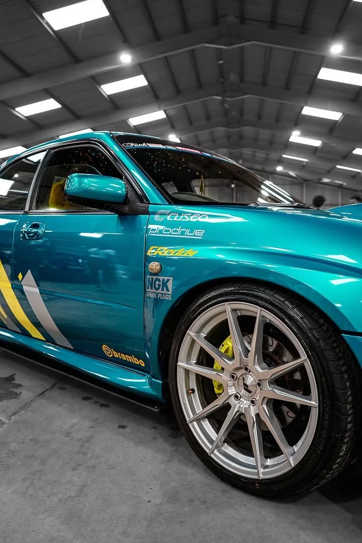 A turquoise sports car with racing decals and large alloy wheels inside a well-lit indoor facility.