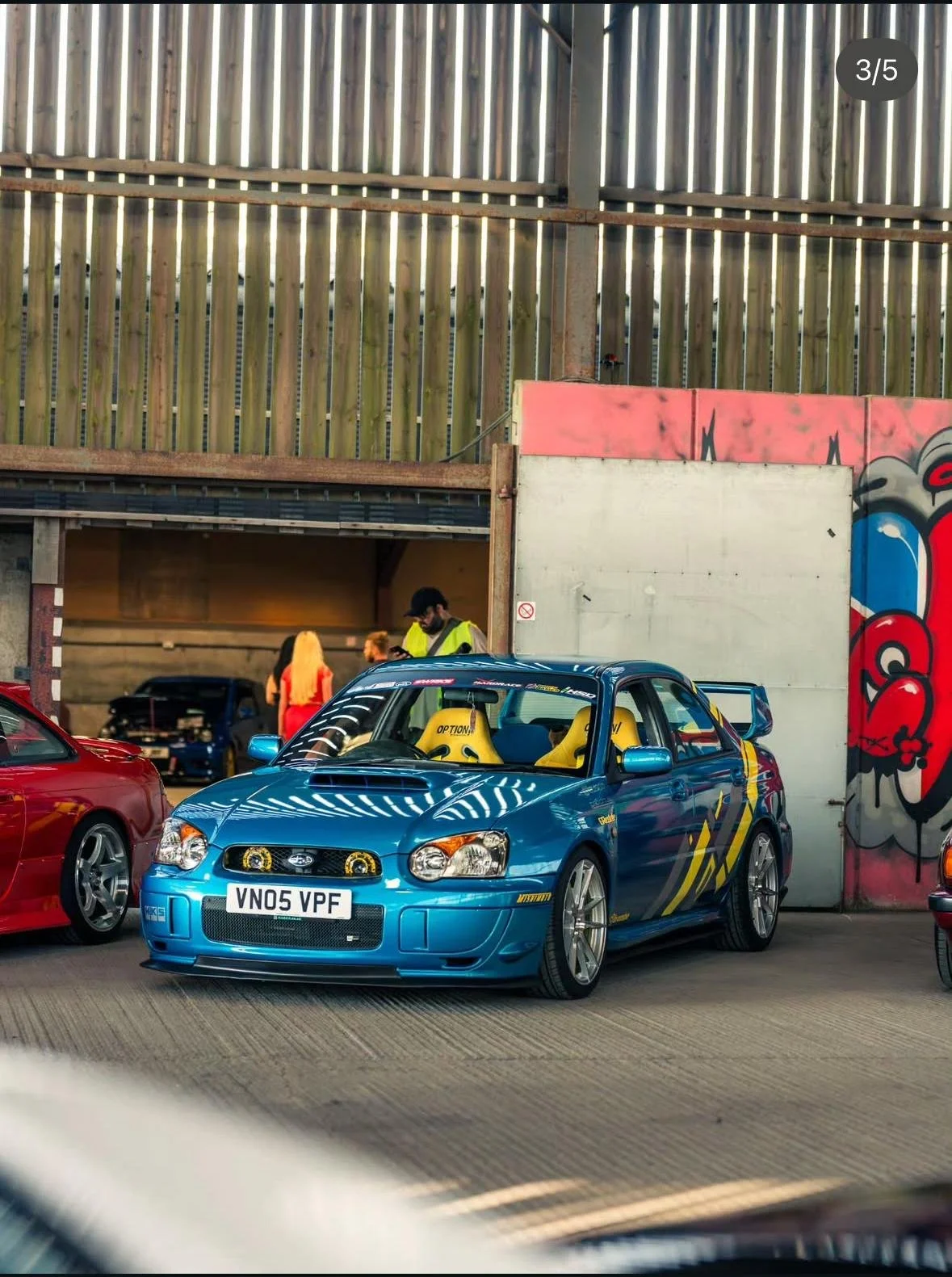 A blue modified hatchback car with yellow racing seats parked in an urban area near a graffiti-covered wall, with two people in the background near a wooden fence.