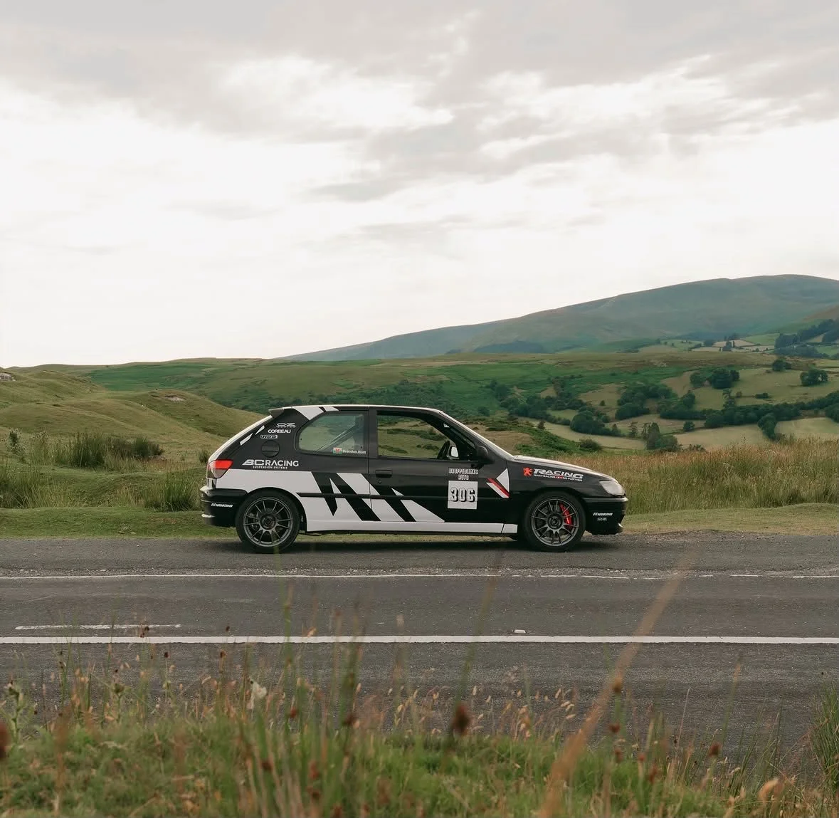 A black and white rally car with racing decals parked on a rural road with green rolling hills and mountains in the background under a cloudy sky.
