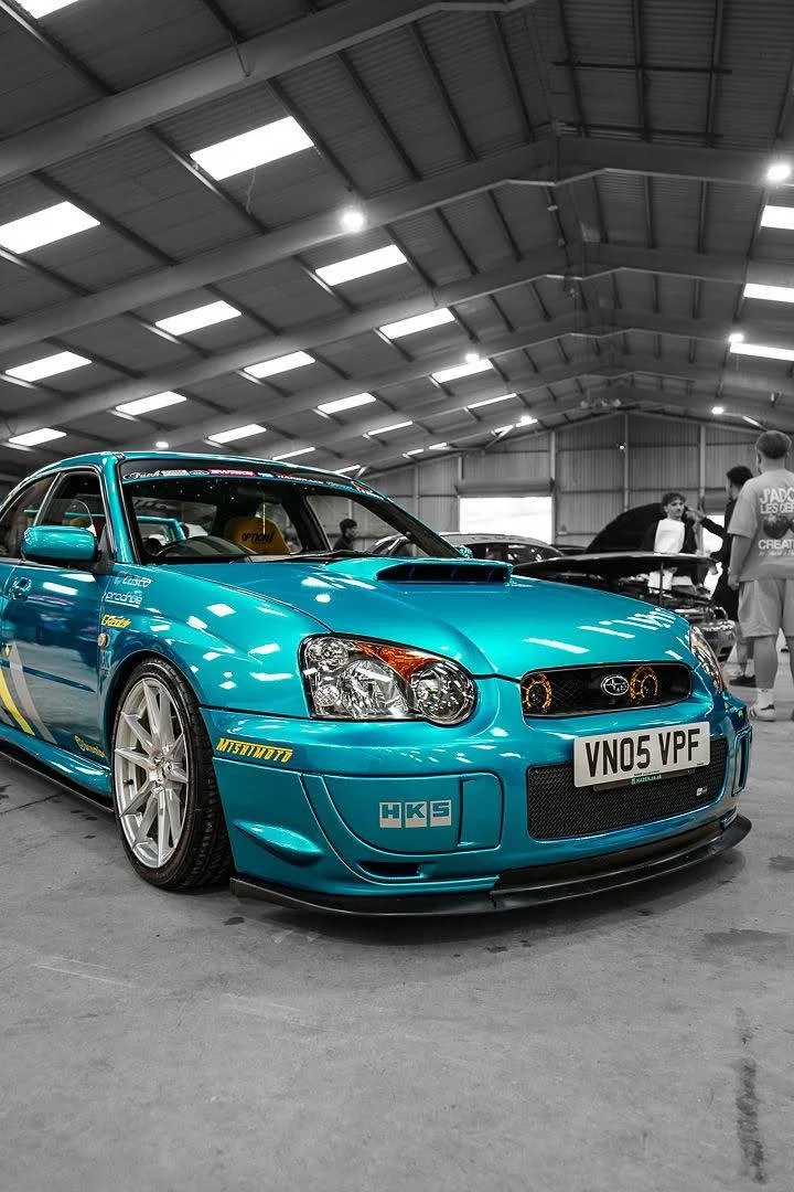 A blue Subaru Impreza WRX STI with yellow decals is on display in a large indoor warehouse at a car show. The car is customized with aftermarket parts, and there are several people in the background admiring other vehicles.