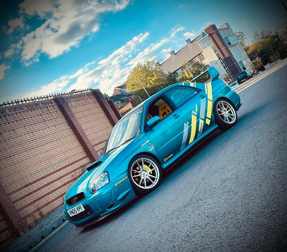A blue race car with yellow and grey decals parked on a street near a brick wall and residential buildings under a partly cloudy sky.