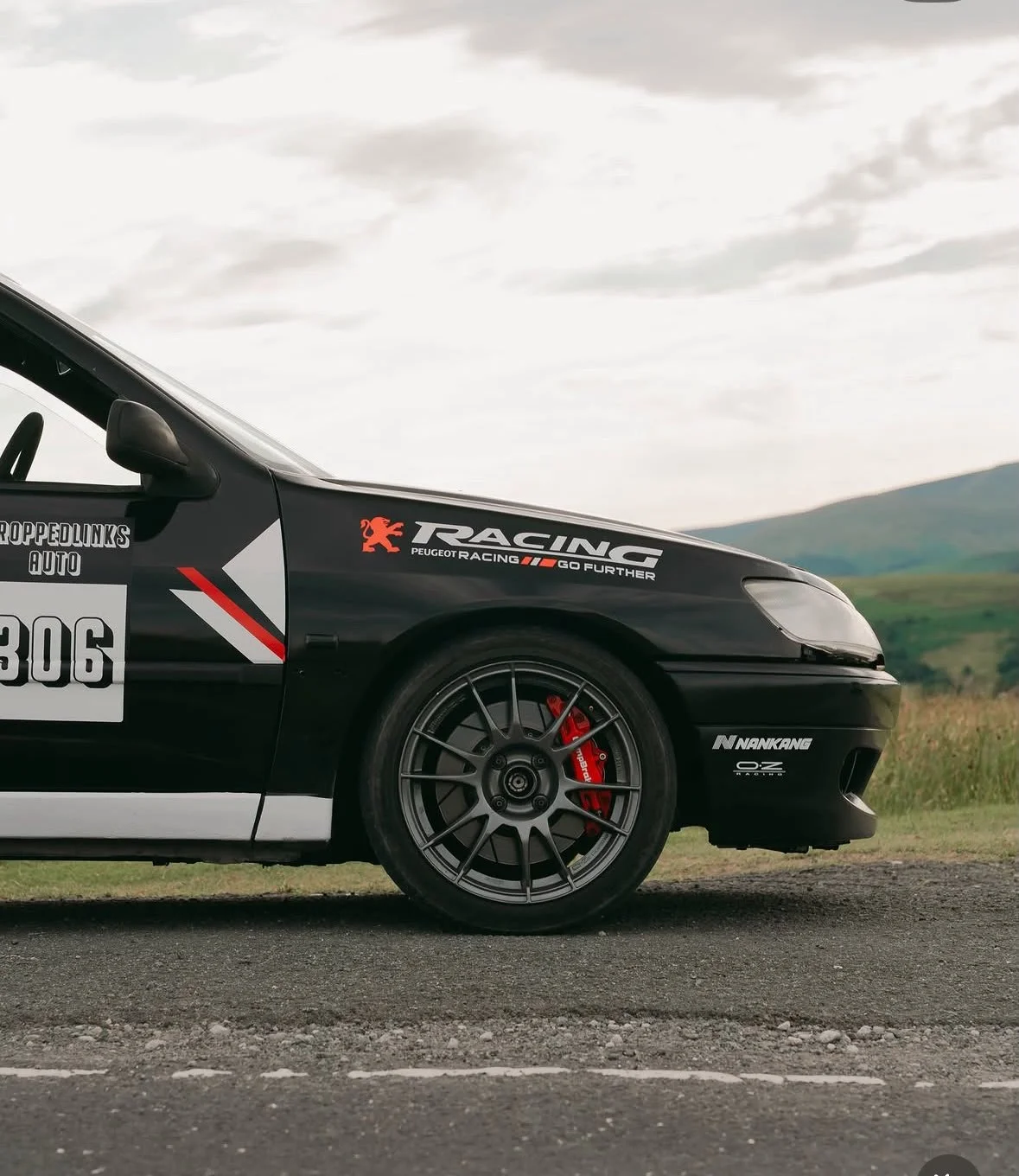Close-up of a black race car parked on the side of a road with green hills and cloudy sky in the background.