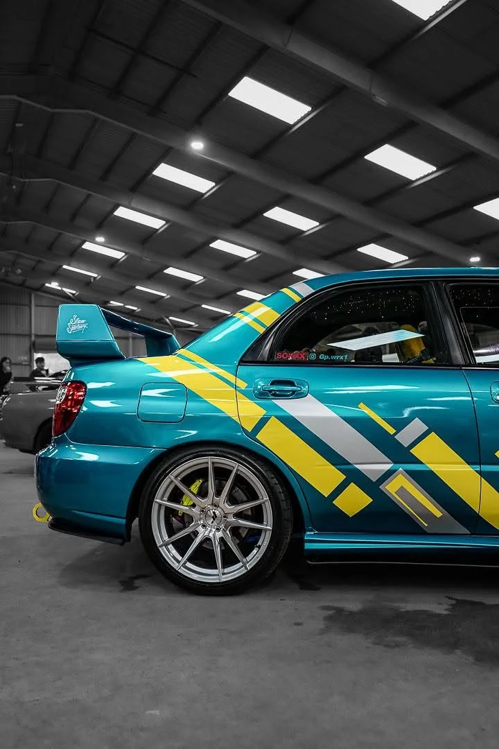 A blue race car with yellow and gray decals in an indoor garage, showing the rear and side.