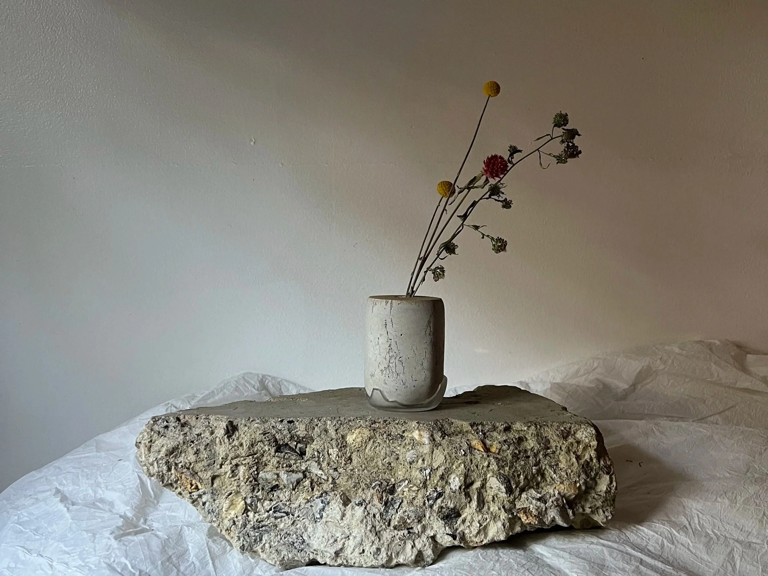 A small vase with dried flowers placed on a large textured rock, with a plain neutral-colored wall in the background.