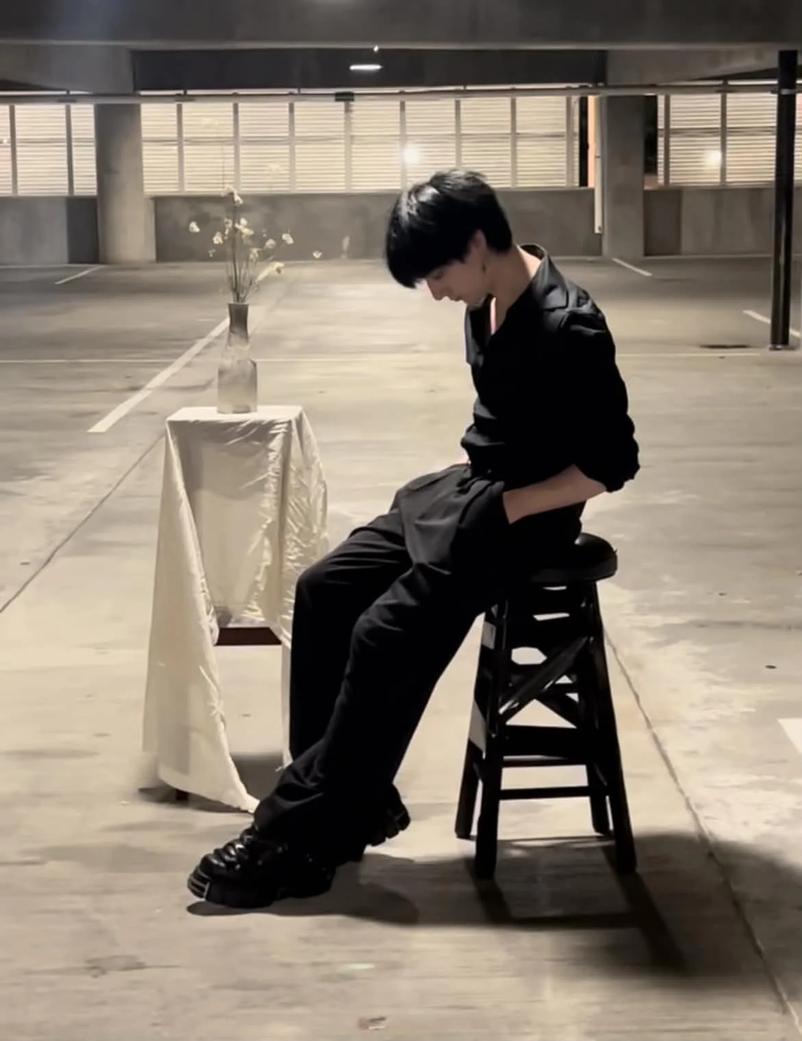 A young man with black hair sitting on a stool in a parking garage, head bowed, hands in pockets, beside a small table with a vase of flowers.