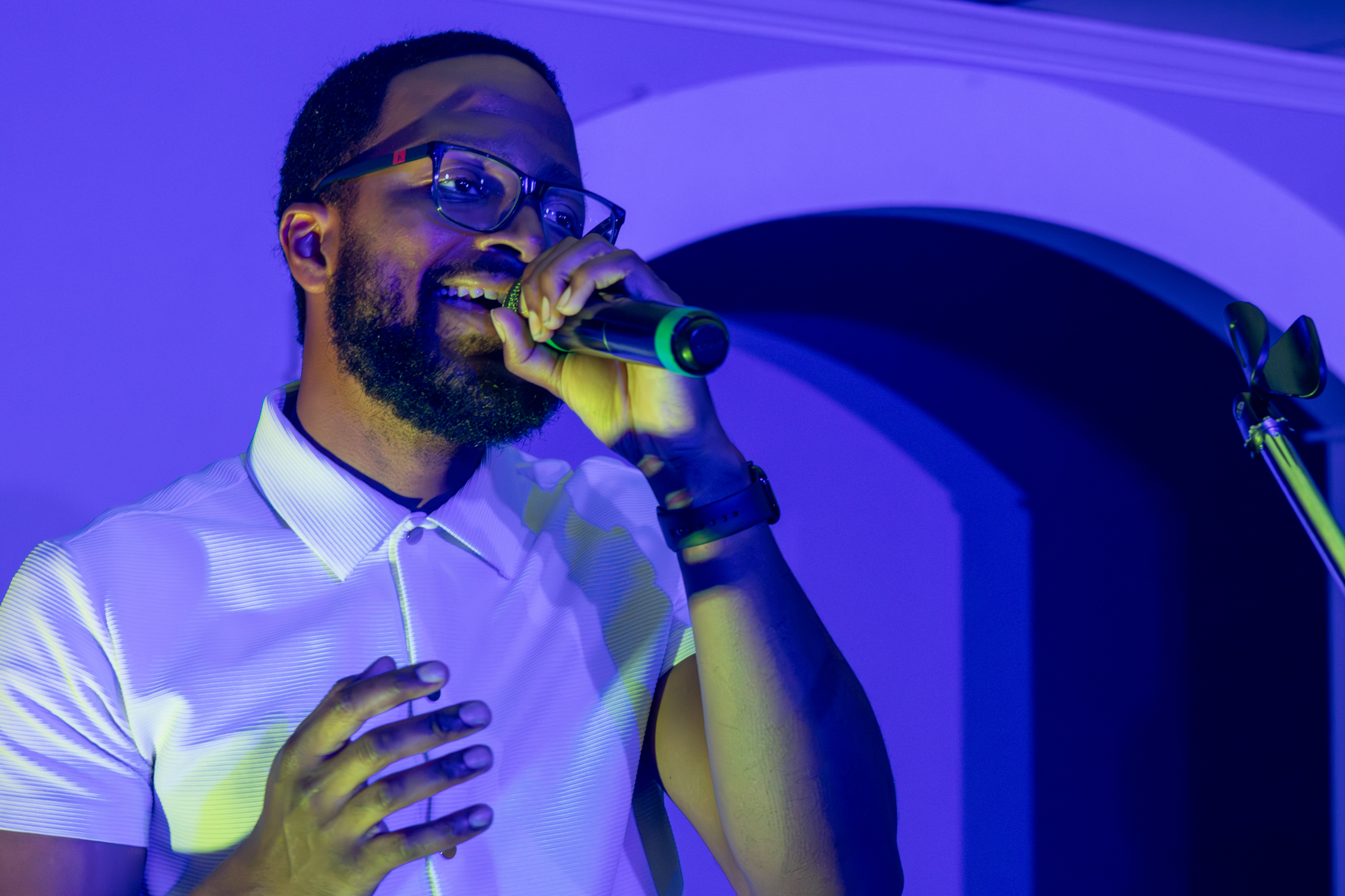 A man wearing glasses and a white shirt singing into a microphone with blue and purple lighting.