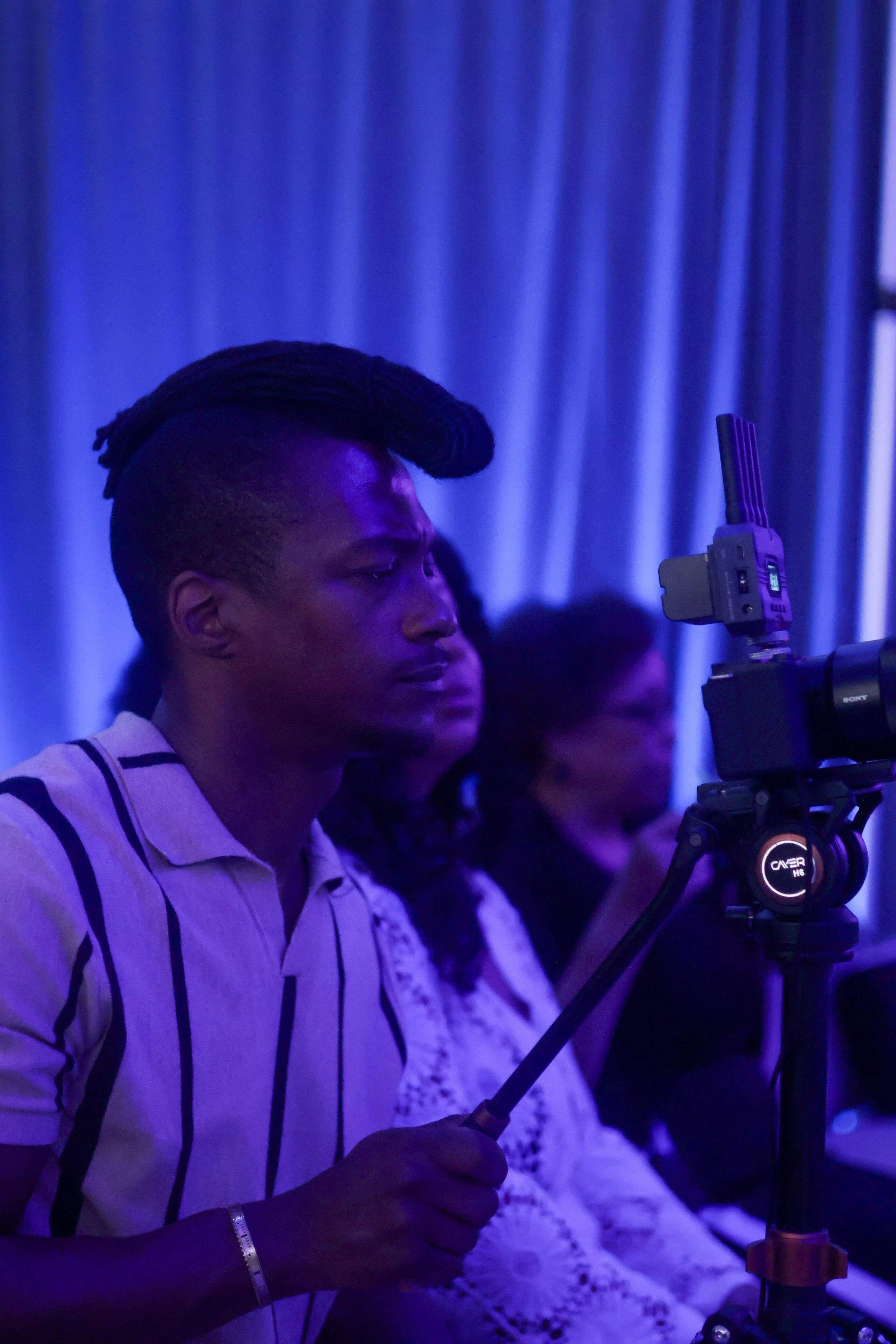A person with a styled haircut and wearing a striped shirt is operating a camera on a tripod in a dimly lit room with blue lighting and curtains in the background.