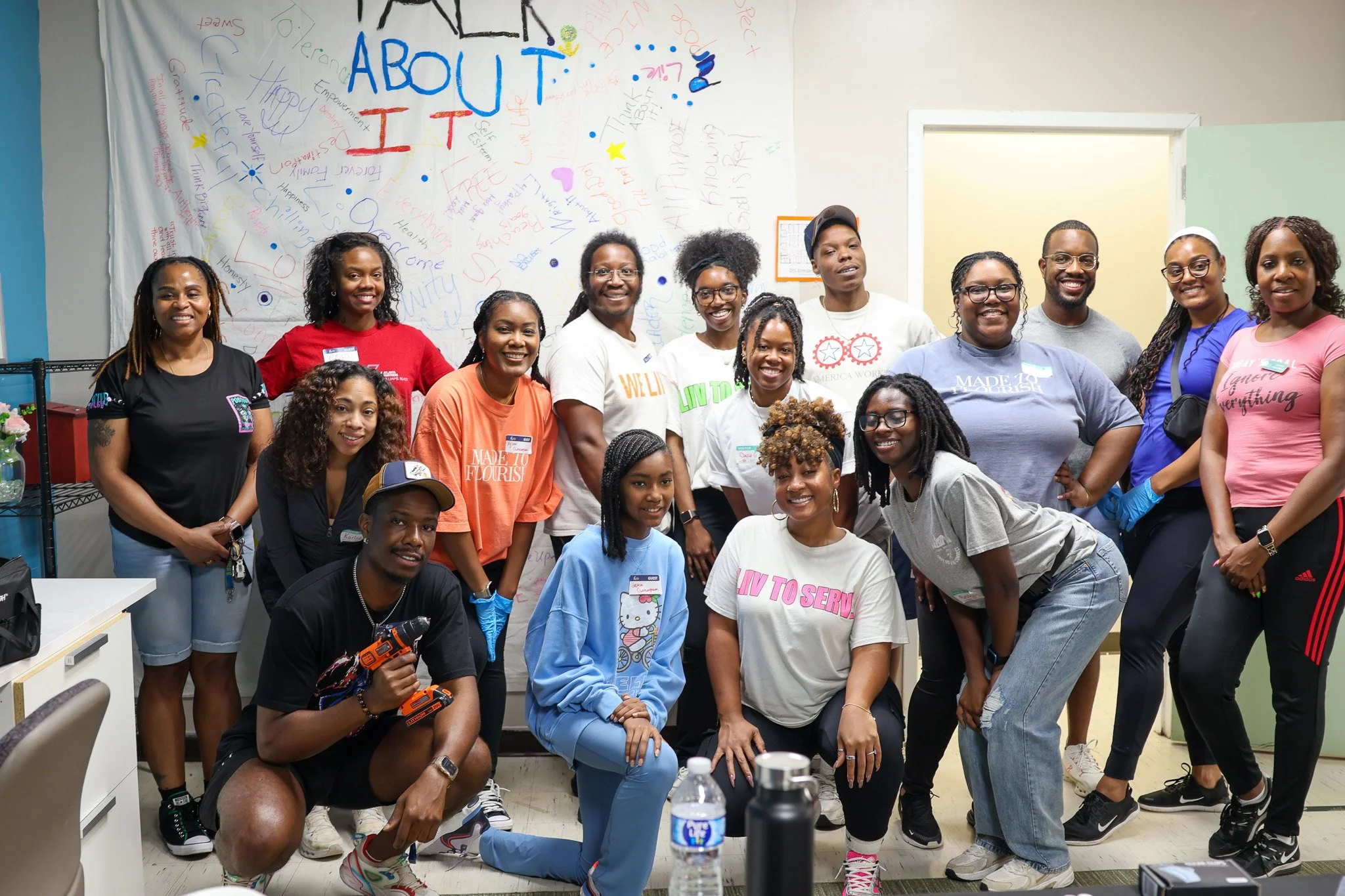 A diverse group of people posing together in a room with a whiteboard behind them, which has colorful handwritten messages and the words 'ABOUT IT' written prominently. There are around fifteen individuals, most smiling and casually dressed.