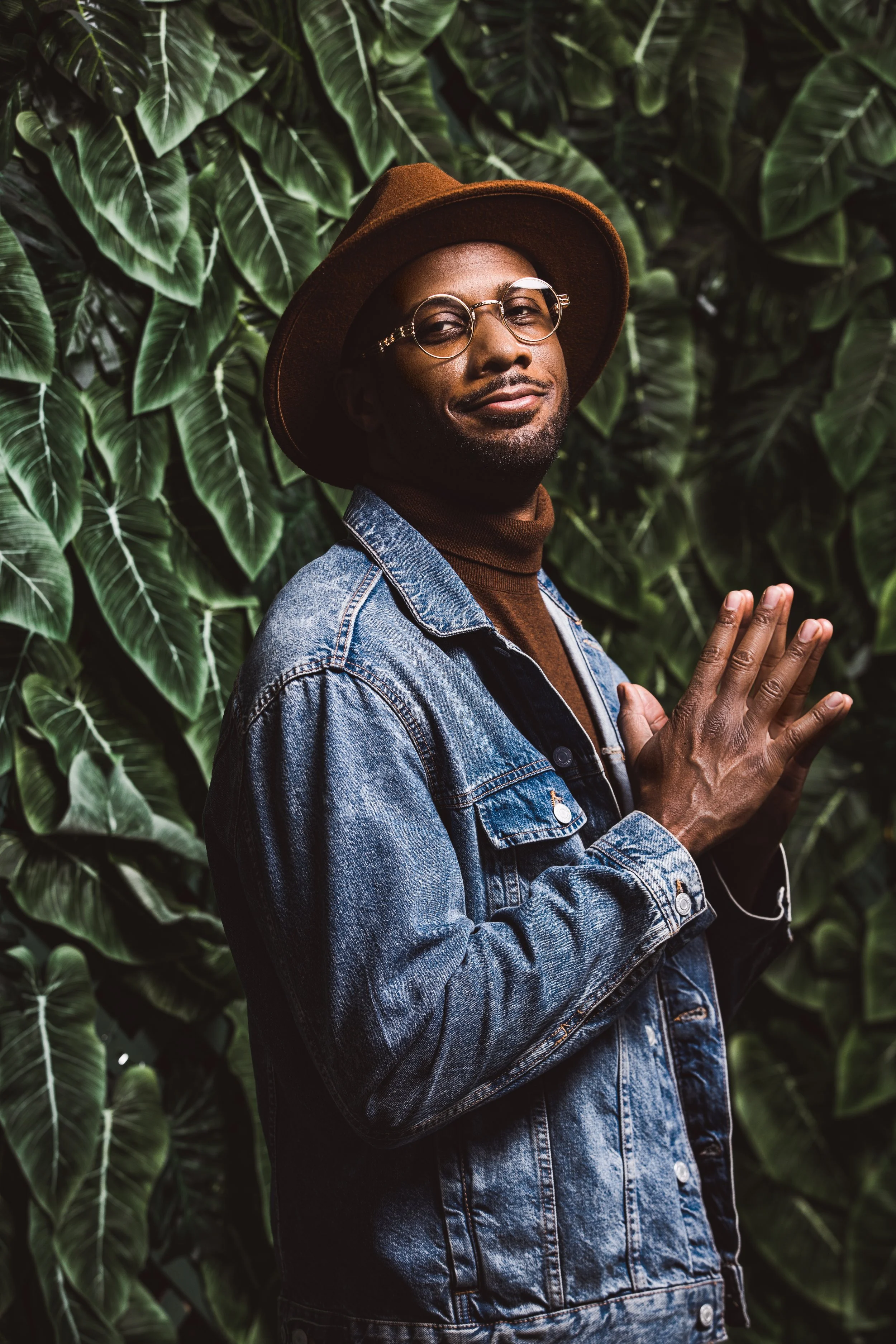 A stylish man wearing glasses, a brown hat, a brown turtleneck, and a denim jacket standing with his hands together in a prayer-like pose in front of lush green leaves.