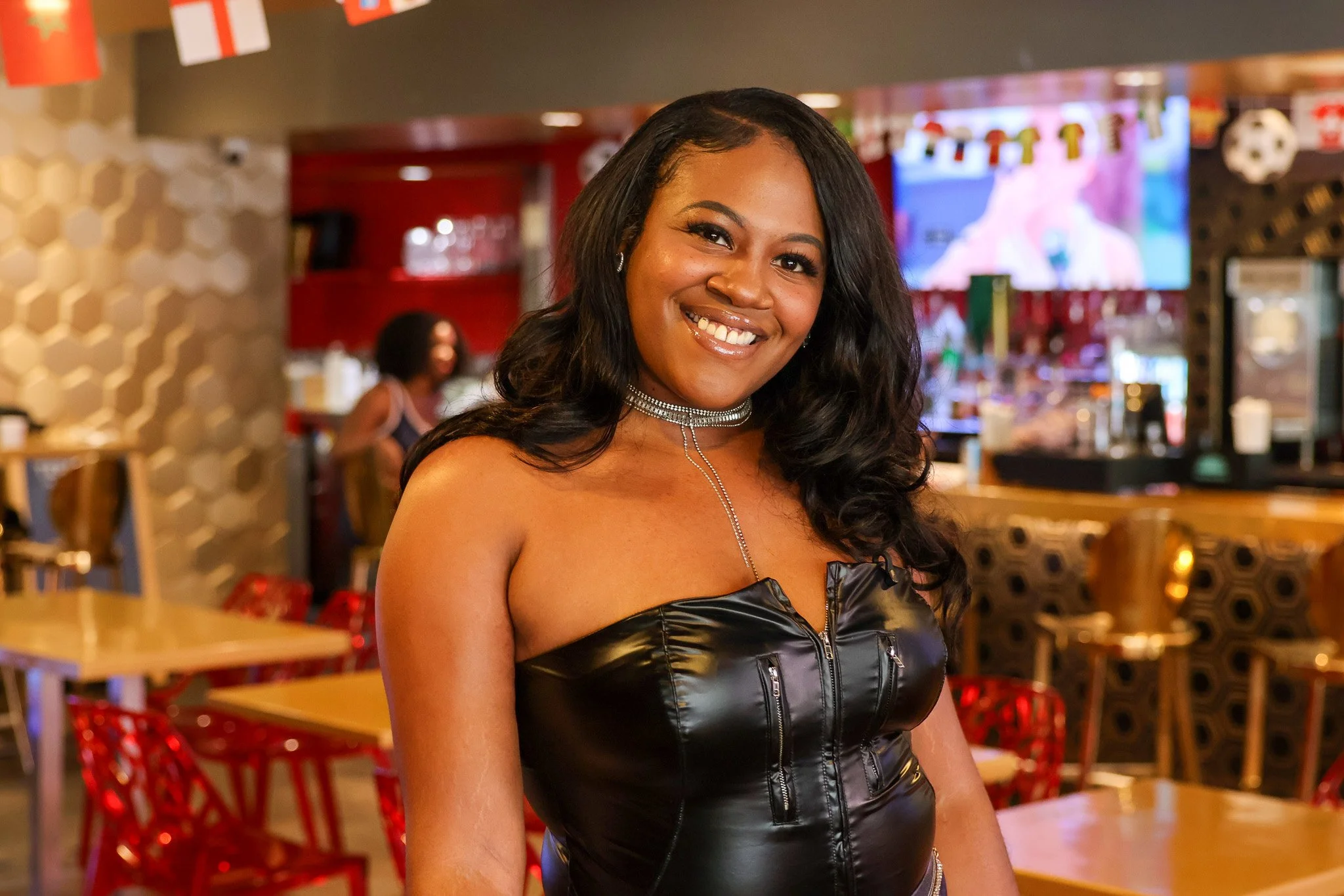 A smiling woman with dark hair and dark eyes, wearing a black leather zip-up top and sparkly jewelry, standing in a decorated restaurant or bar area with red chairs and a TV screen in the background.