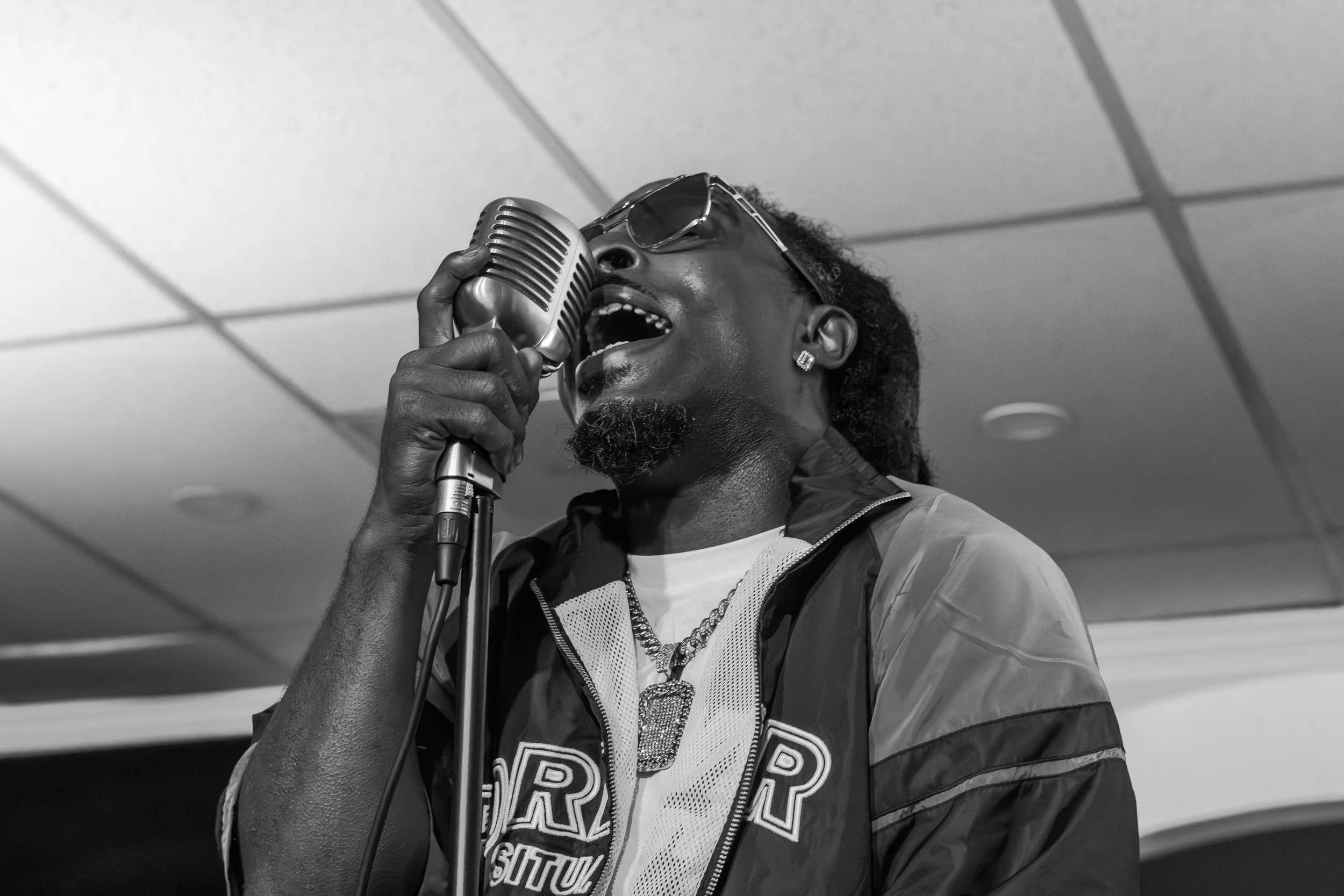 A Black man singing into a vintage microphone, wearing sunglasses and a leather jacket, in an indoor setting.