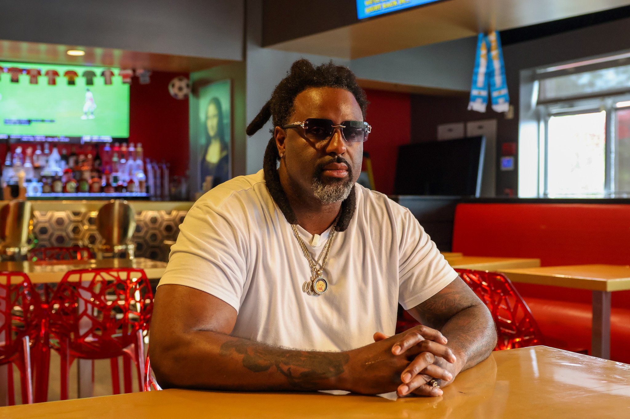 A man with dreadlocks and sunglasses sitting at a table in a restaurant or cafe, with colorful chairs and a TV screen in the background.