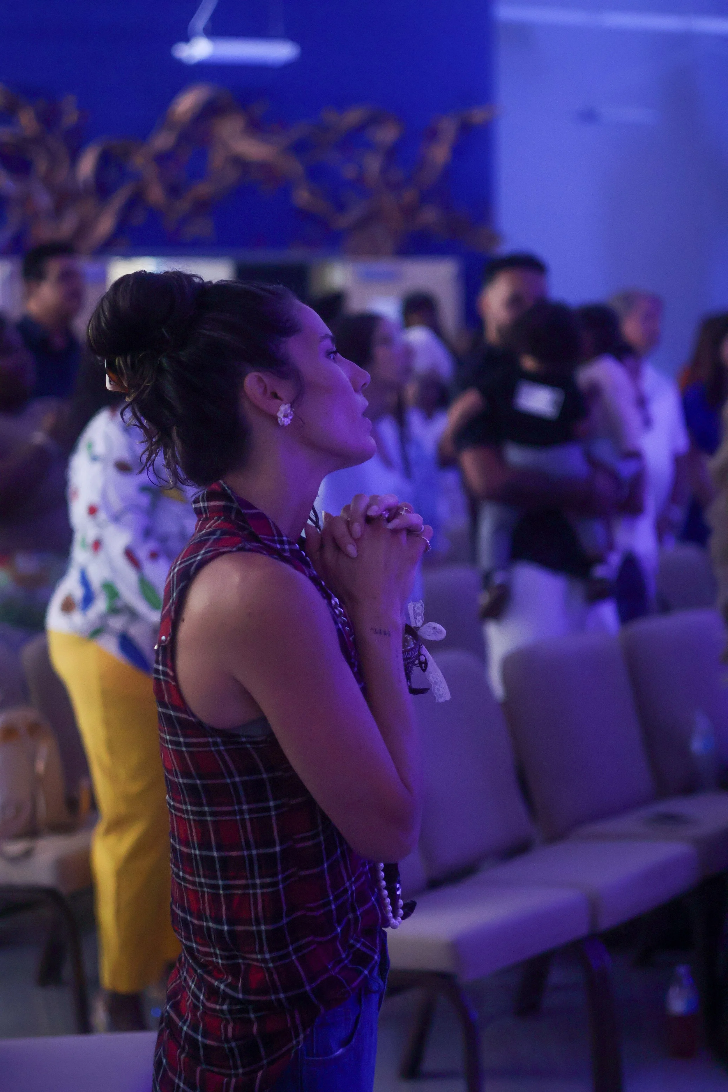 A woman with dark hair styled in an updo, wearing earrings and a plaid sleeveless dress, is standing with her hands clasped and closed eyes in a dimly lit setting, surrounded by other people, some with their hands also clasped.