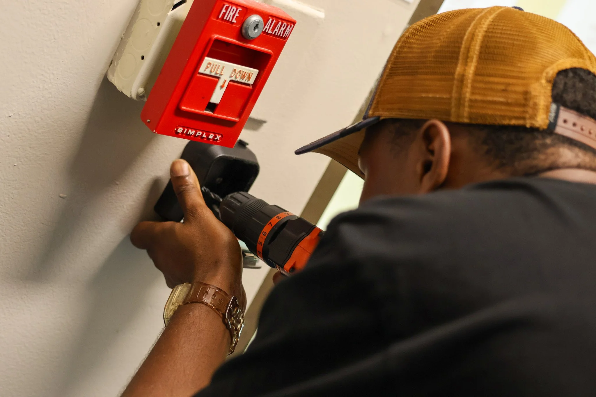 A person wearing a yellow baseball cap working on a fire alarm system using a cordless drill, with a red fire alarm box mounted on a wall nearby.