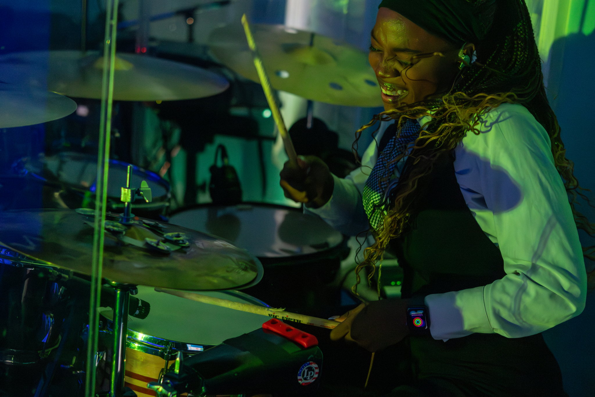 A woman with braided hair and glasses, wearing a white shirt, is playing drums and smiling in a dimly lit room with green lighting.
