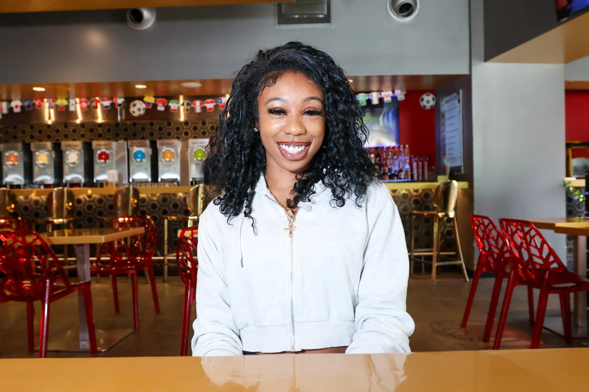 A woman with curly black hair smiling while sitting at a table in a restaurant with colorful chairs and sports-themed decorations in the background.