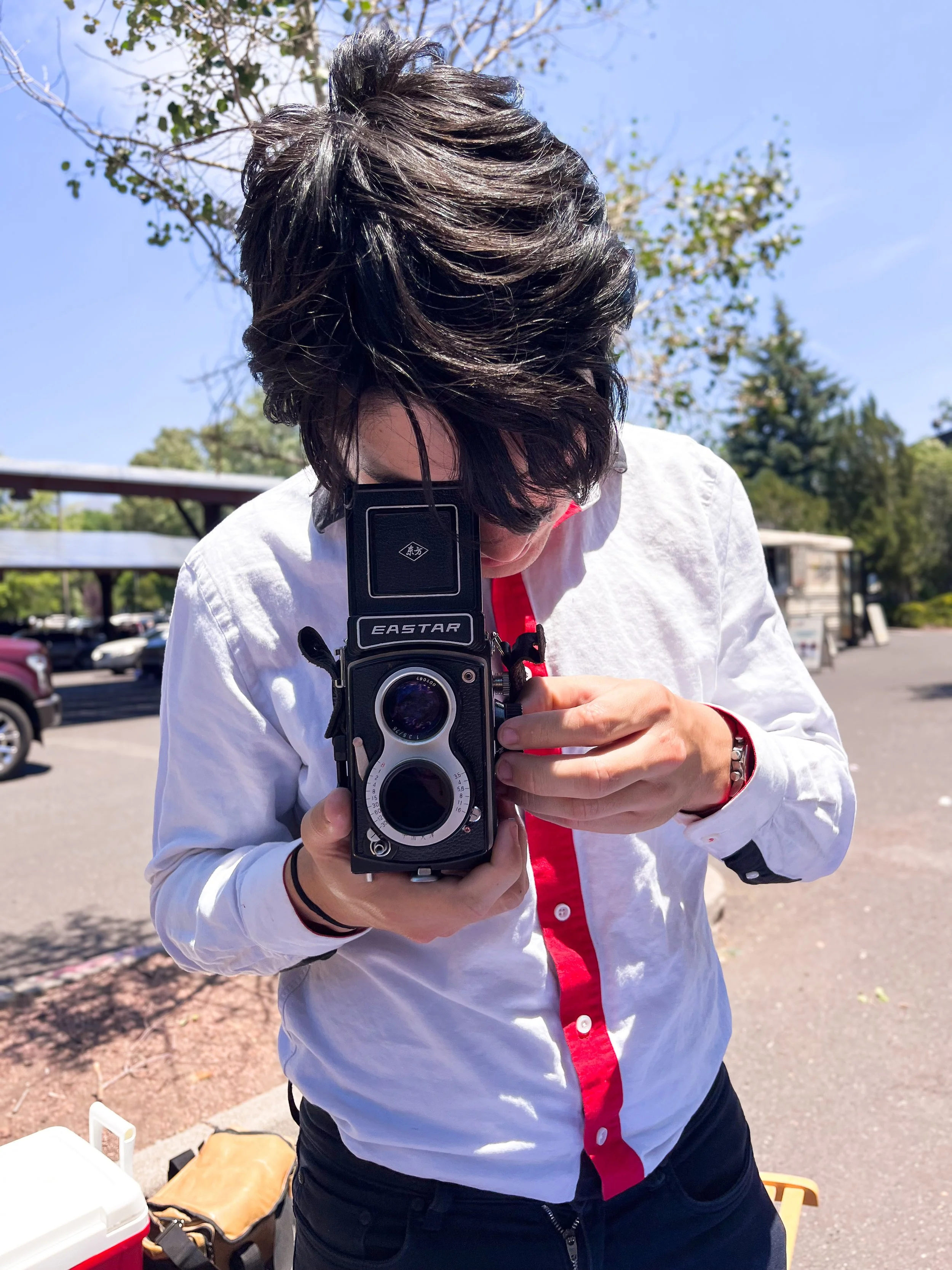 Person with dark, wavy hair taking a photo with a vintage twin-lens reflex camera outdoors on a sunny day, with trees and a parking lot in the background.