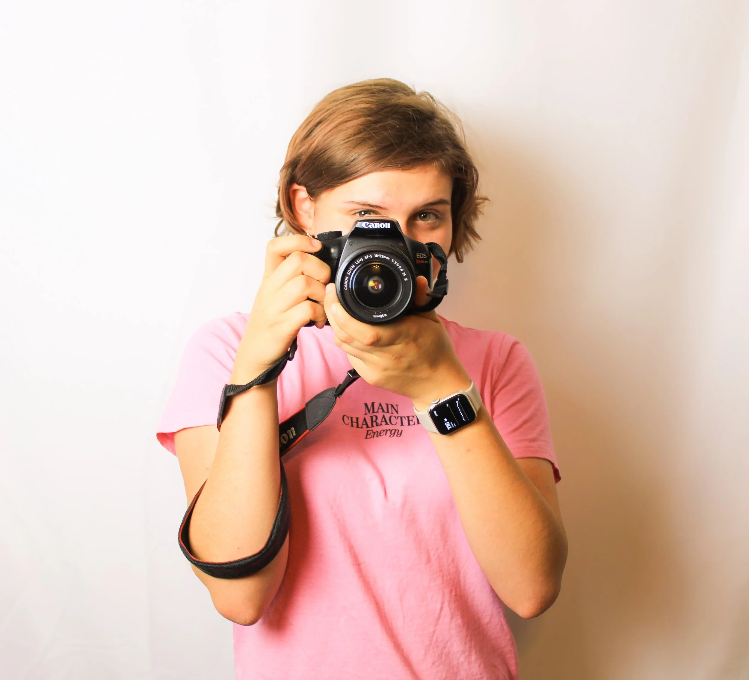 Woman with short brown hair holding a Canon camera in front of her face, wearing a pink shirt, and a smartwatch, against a plain white background.