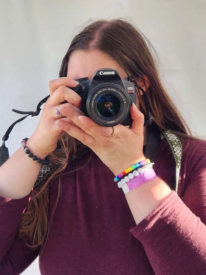 Person taking a photo with a Canon camera, partially obscuring their face, wearing bracelets and a maroon shirt.