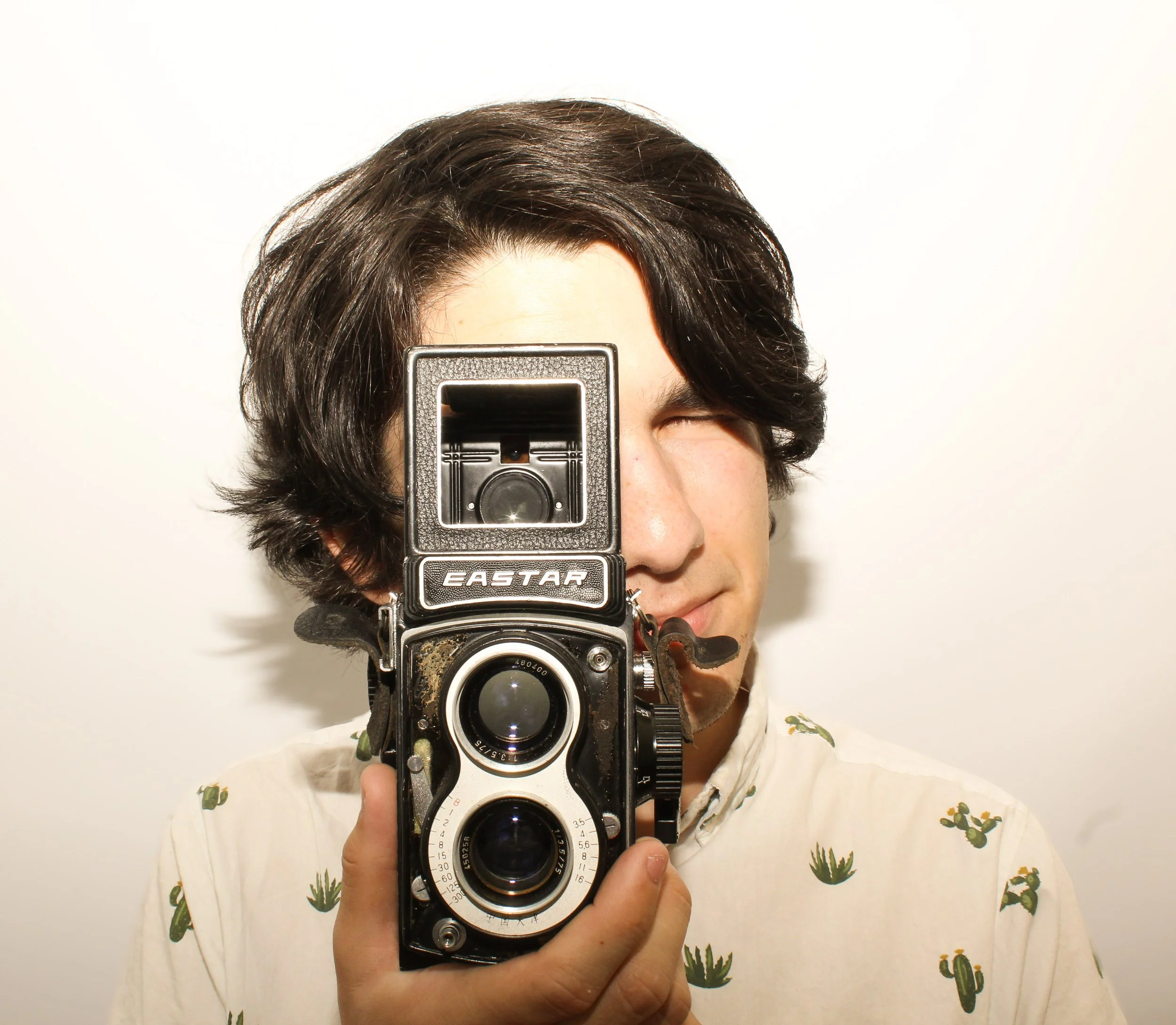 Person taking a selfie using a vintage camera, with dark wavy hair, partially visible face, against a plain white background, wearing a shirt with cactus patterns.
Located in Mesa, Arizona at Flash Haus
