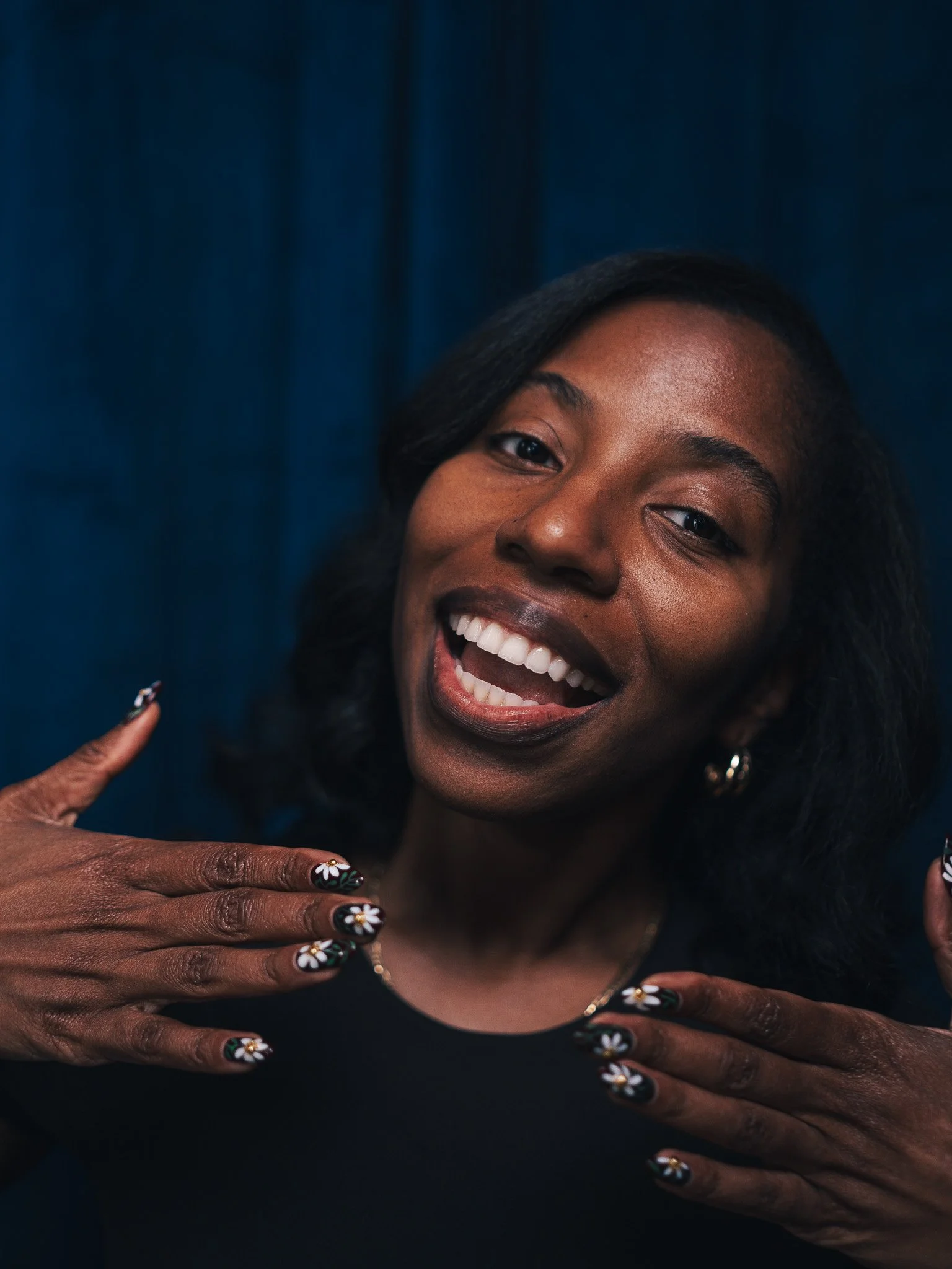 A smiling woman (Michelle Turpin) with dark skin and black hair, showing her teeth, with her hands in front of her chest. Her nails are decorated with maroon nail polish and white daisy designs.