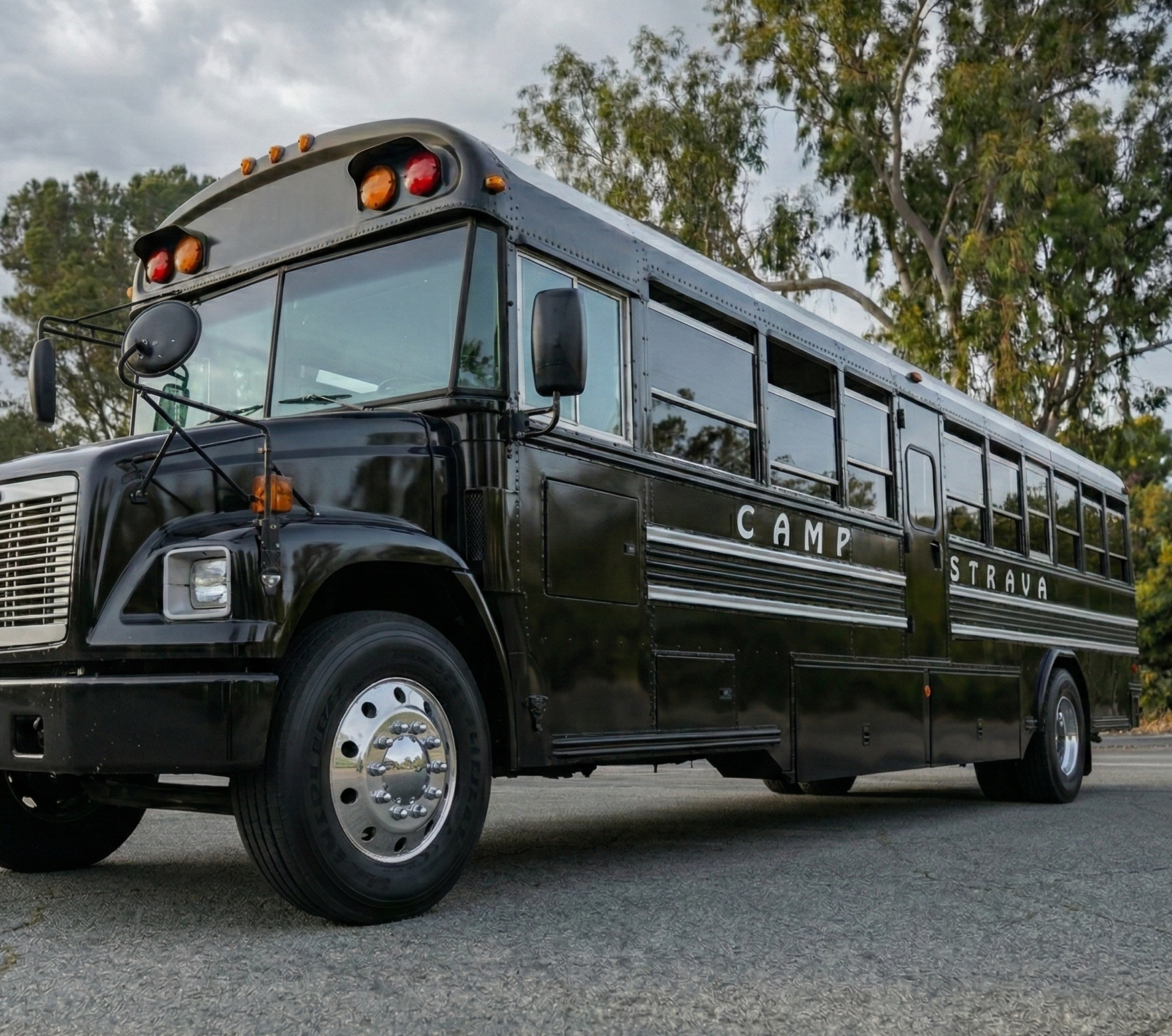 A black school bus with the text 'CAMP STRAVA' on its side, parked outdoors with trees and cloudy sky in the background.