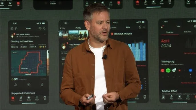 Man standing on stage giving a presentation with three large smartphone screens displaying fitness and workout data in the background.