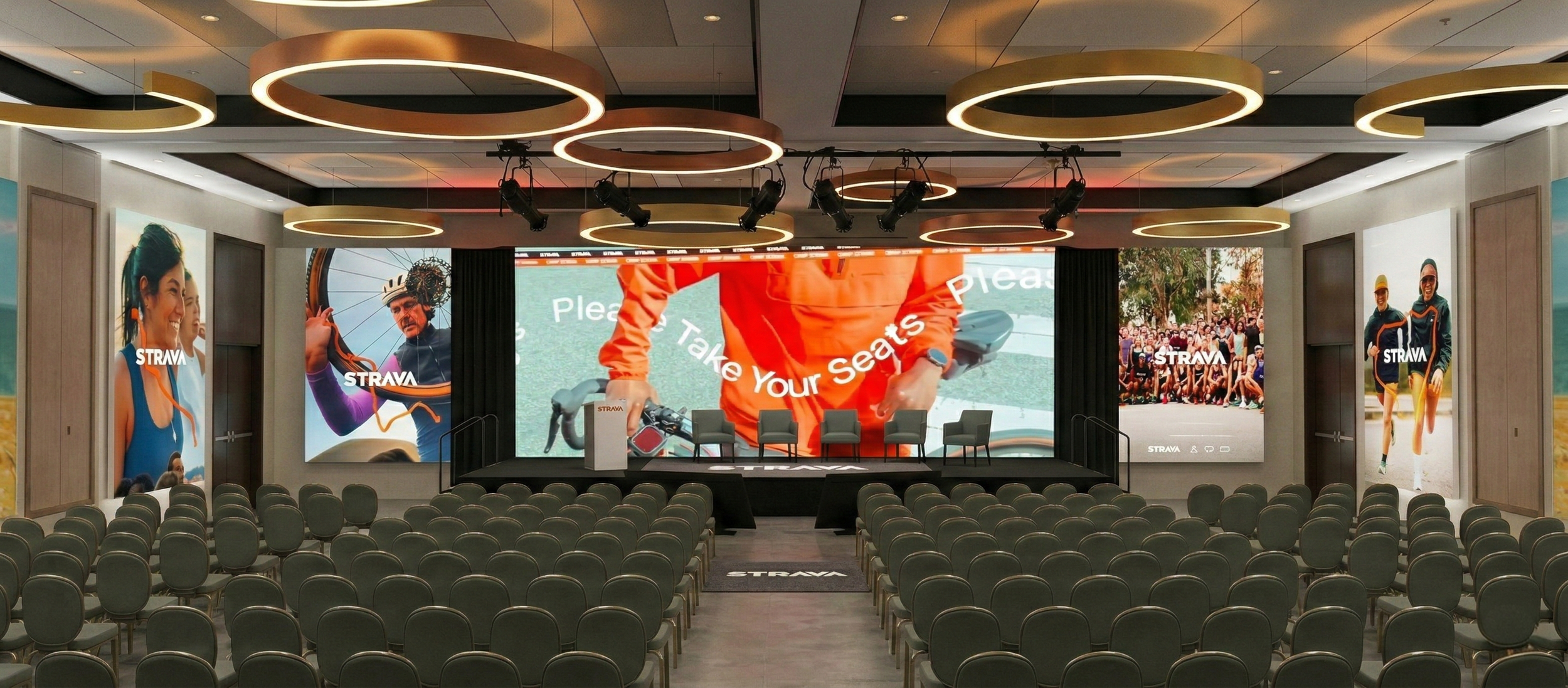 Conference room set up with rows of chairs facing a stage with four chairs, a podium, and large digital screens displaying 'Strava' branding and images of athletes.