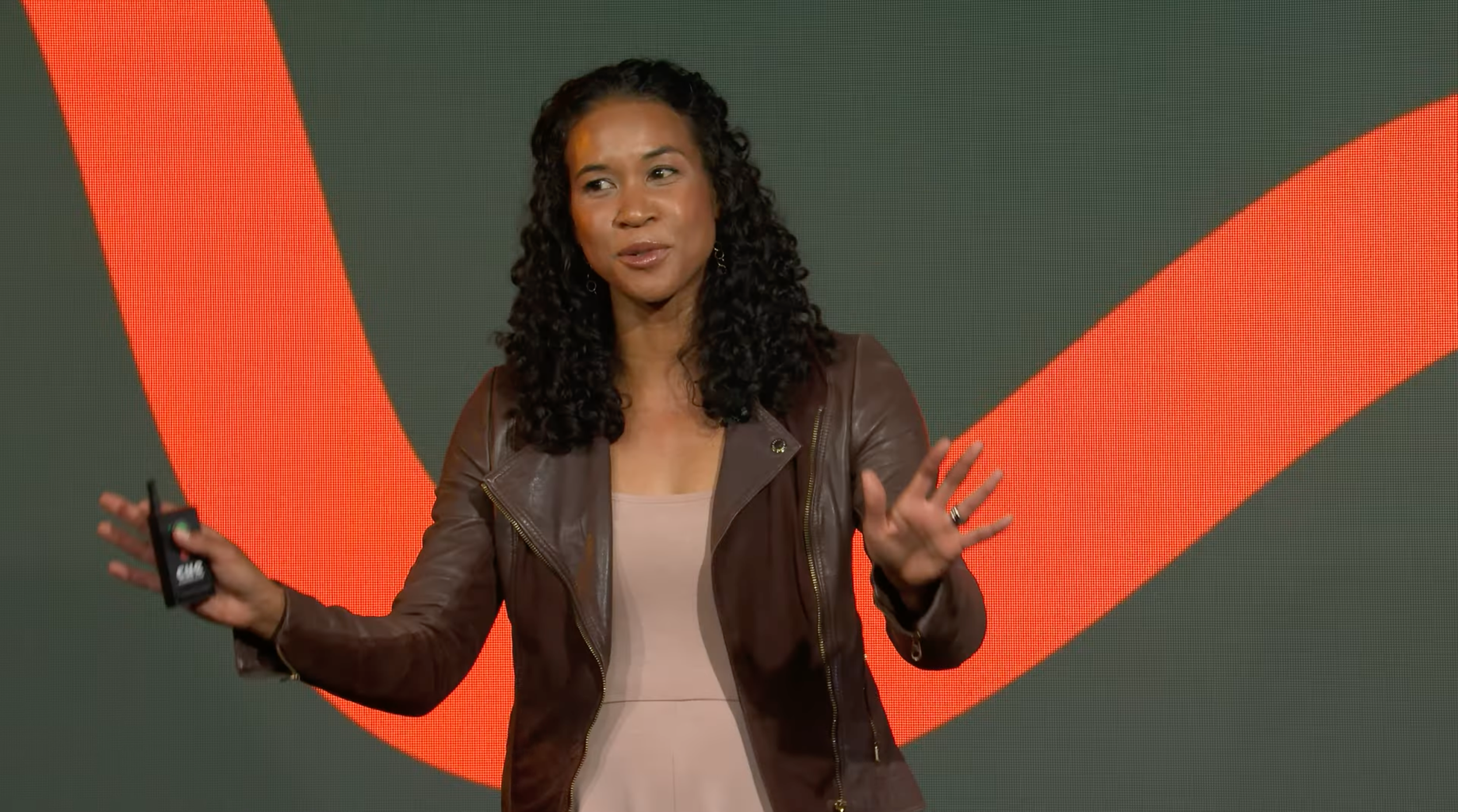 A woman with curly black hair wearing a brown leather jacket and light-colored top, standing on a stage with a large red and green background, speaking and gesturing with her hands.