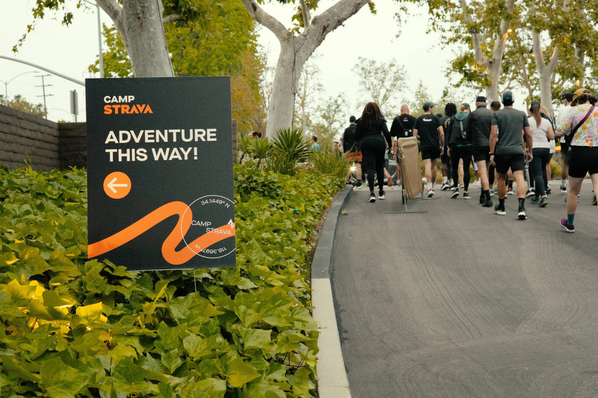 Sign for Camp Strava with an arrow pointing left and a group of people walking on the sidewalk near trees and bushes.