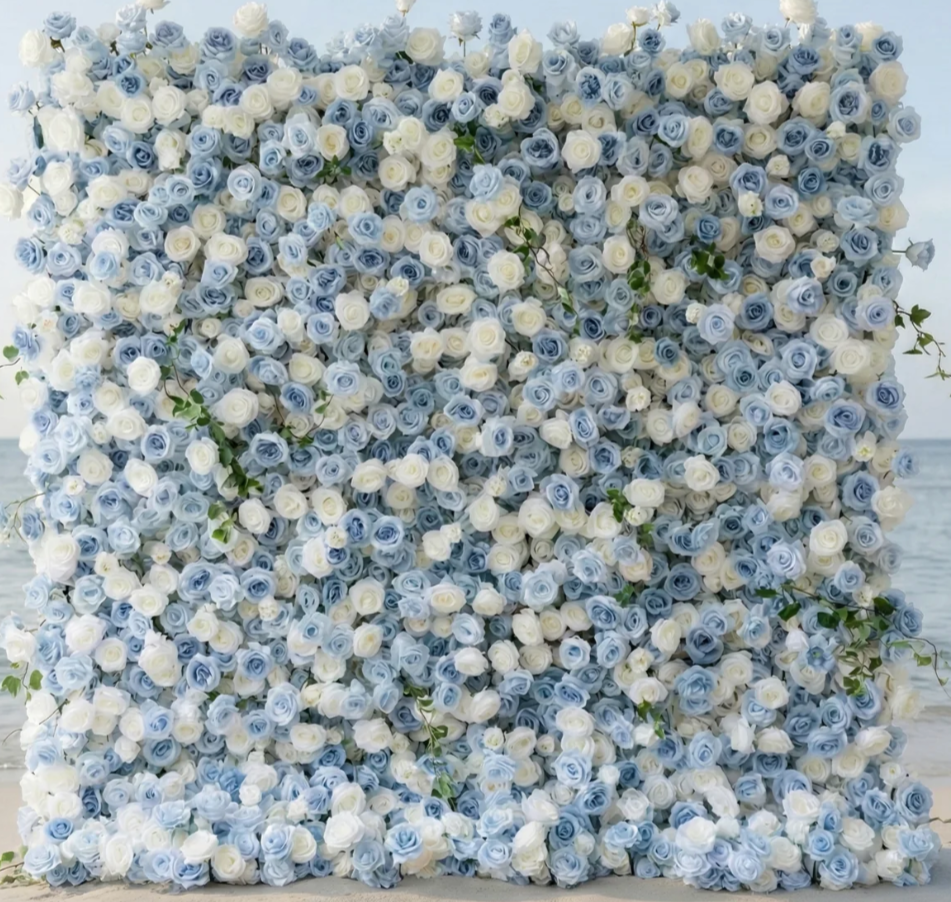A large decorative wall covered in white and light blue roses, with some green leaves, located at a beach or seaside setting.