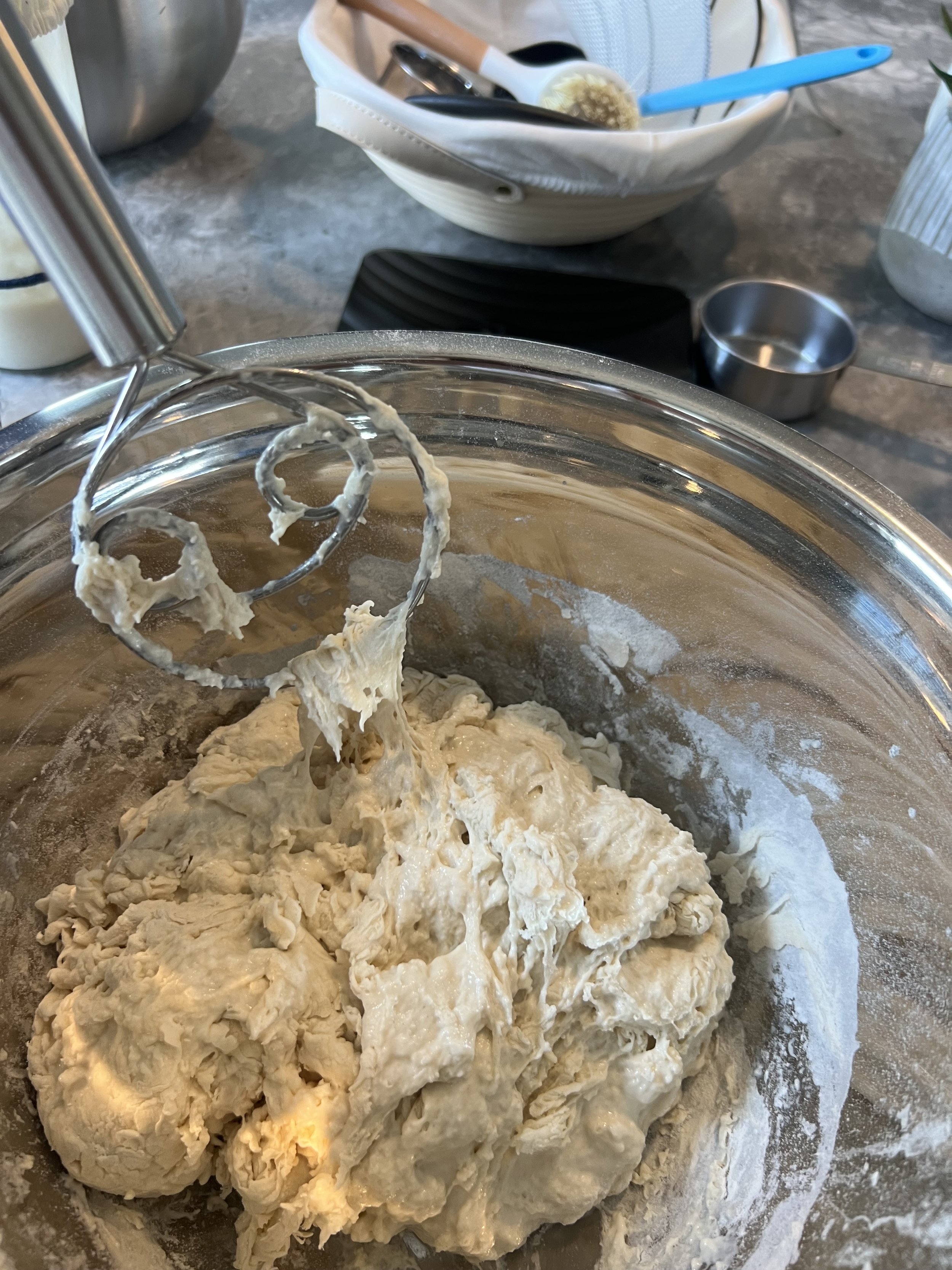 Close-up of dough being mixed in a large metal bowl with a paddle attachment from a stand mixer, with a bowl containing utensils in the background.