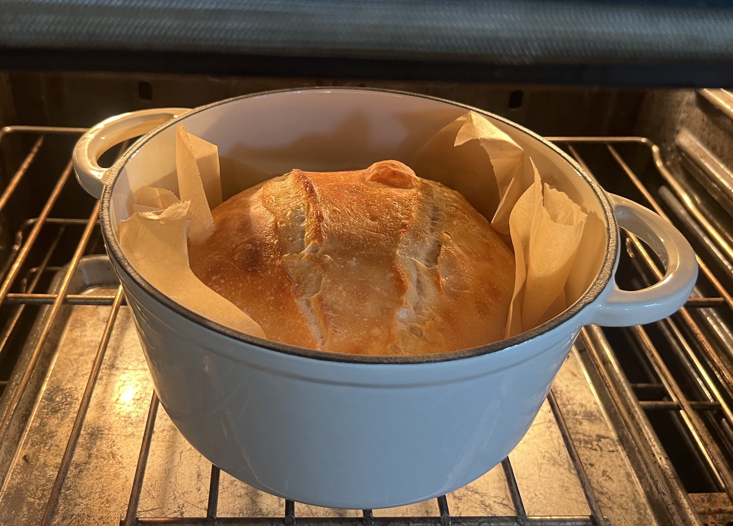 A round loaf of bread baking in an oven inside a ceramic baking dish lined with parchment paper.