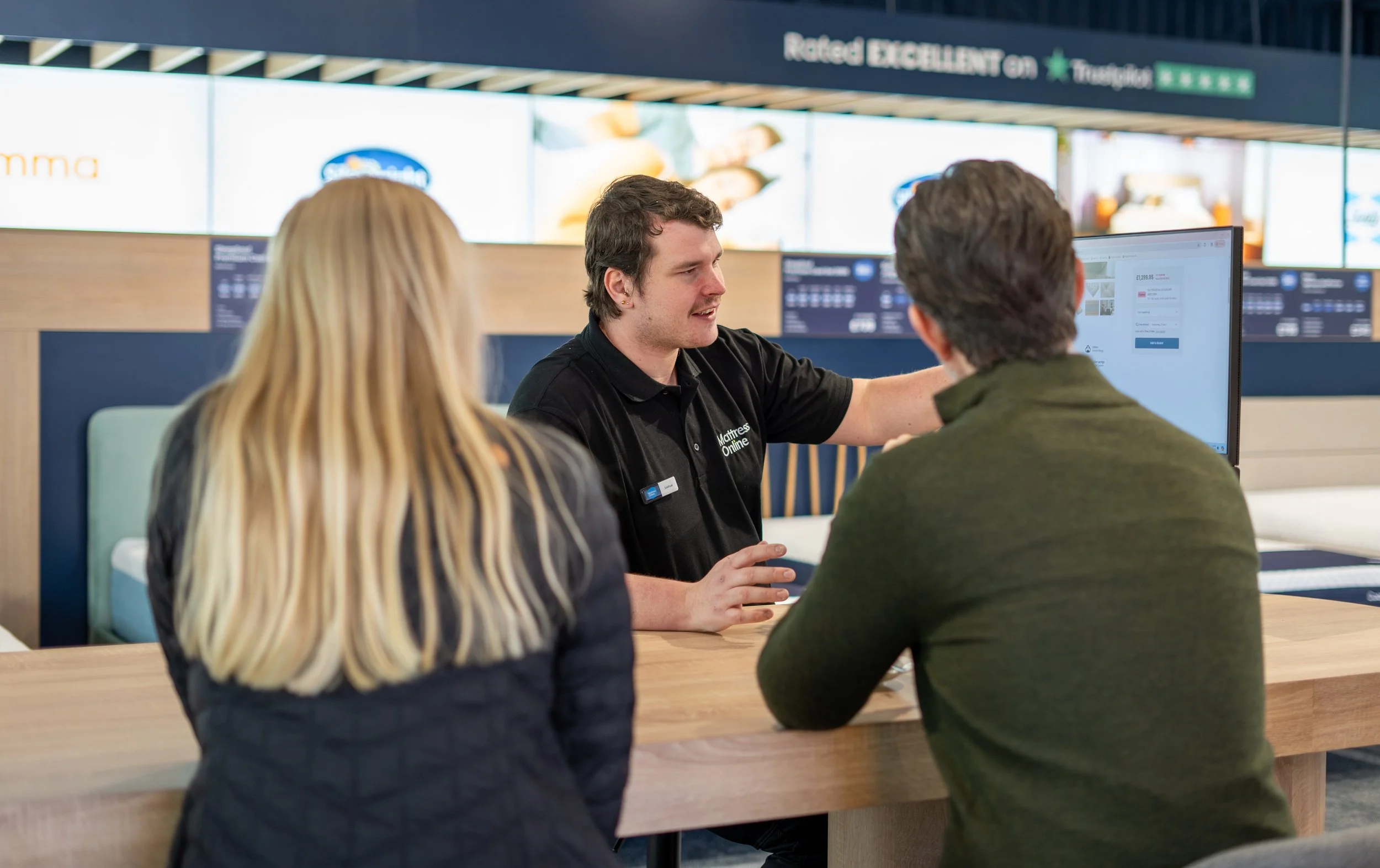 A sales associate is helping two customers at a furniture store counter. The store has digital screens and signage advertising rated high for customer satisfaction.