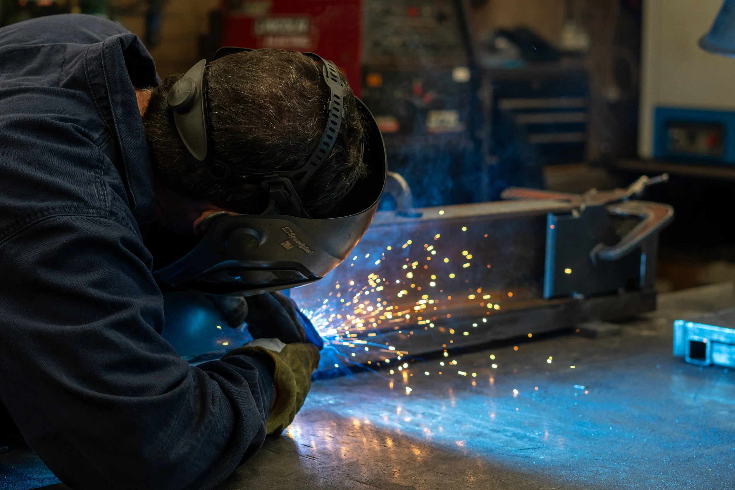 A person welding in a workshop, wearing protective gear including a welding helmet and gloves, sparks flying from the welding process.