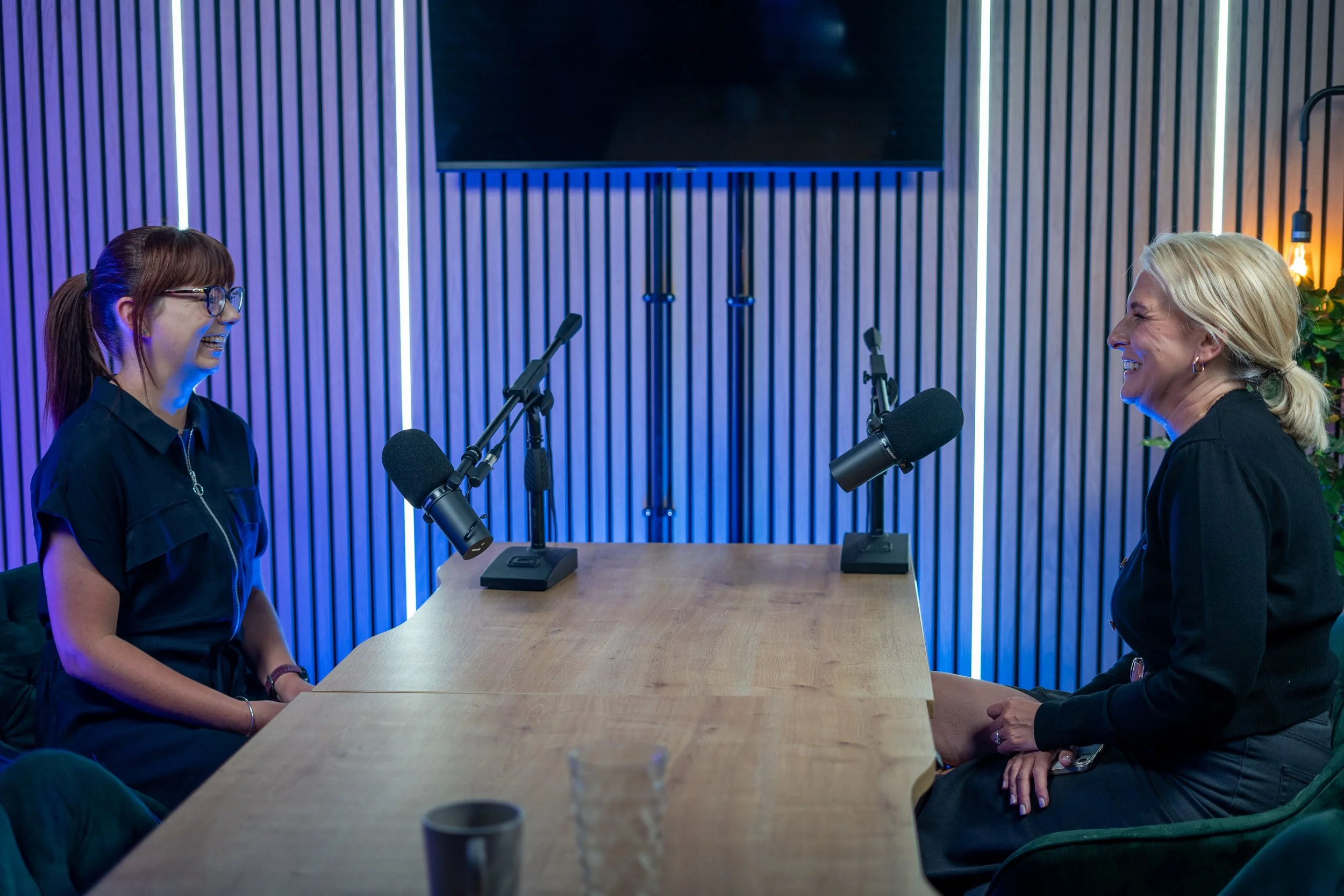 Two women sitting across from each other at a wooden table in a recording studio, smiling and facing each other, with microphones in front of them and a large screen on the wall behind them.