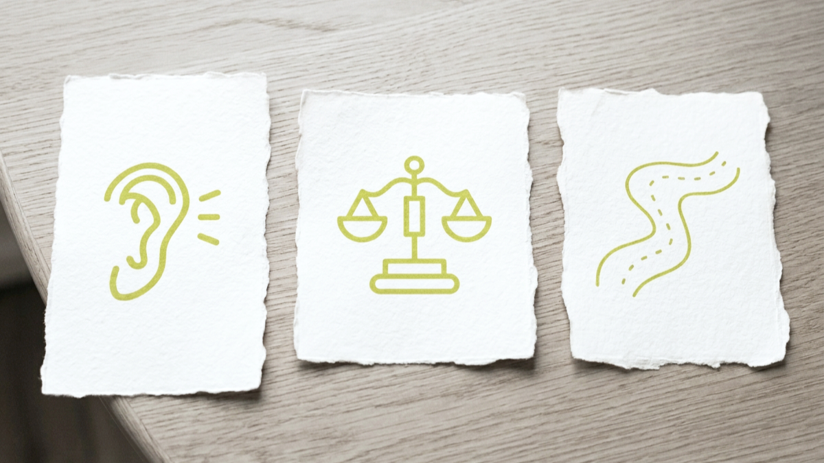 Three pieces of torn white paper with green icons: an ear, a balance scale, and a winding road, on a light wood surface.