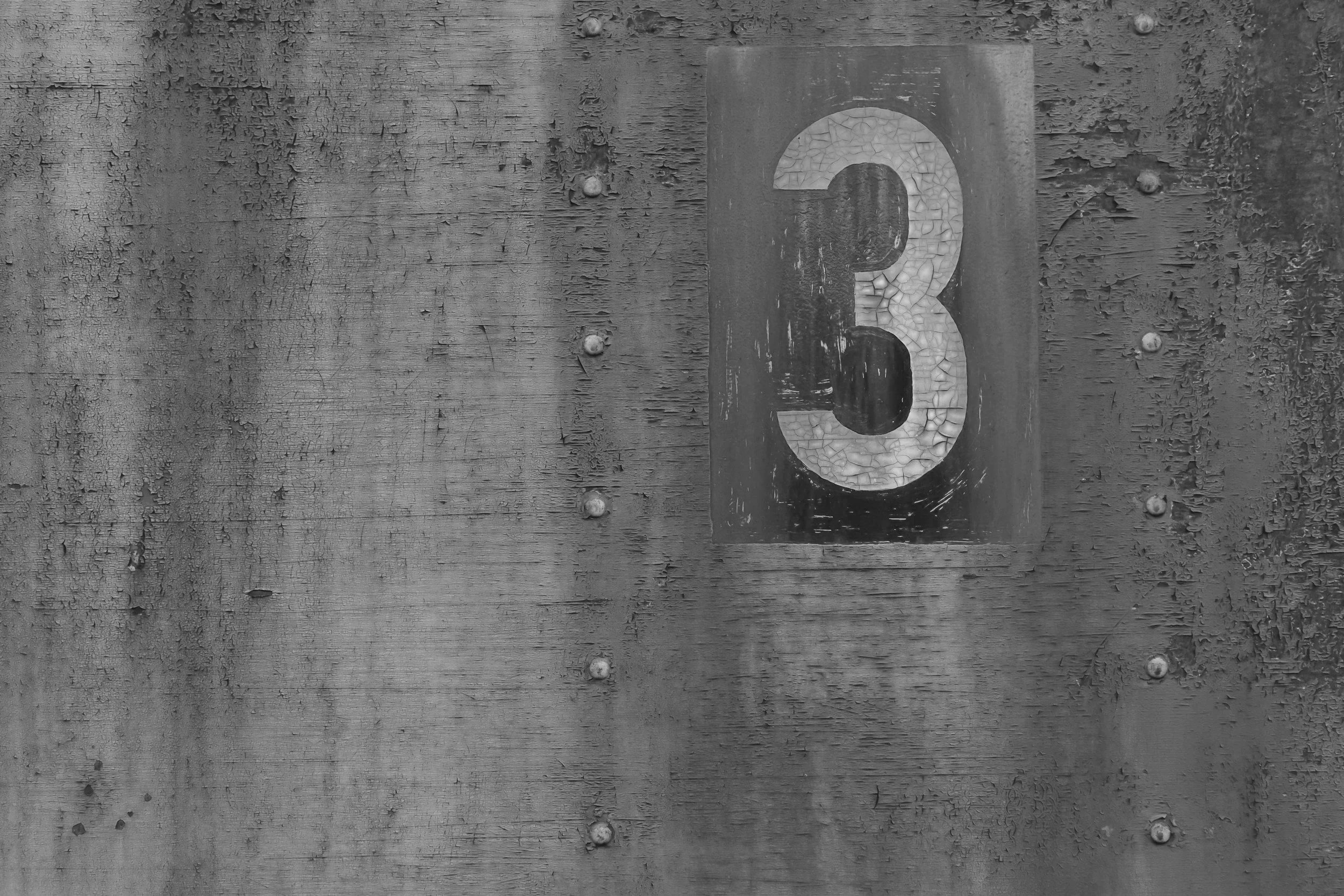 Close-up of an aged wooden surface with a metal number '3' attached, showing peeling paint and rust.