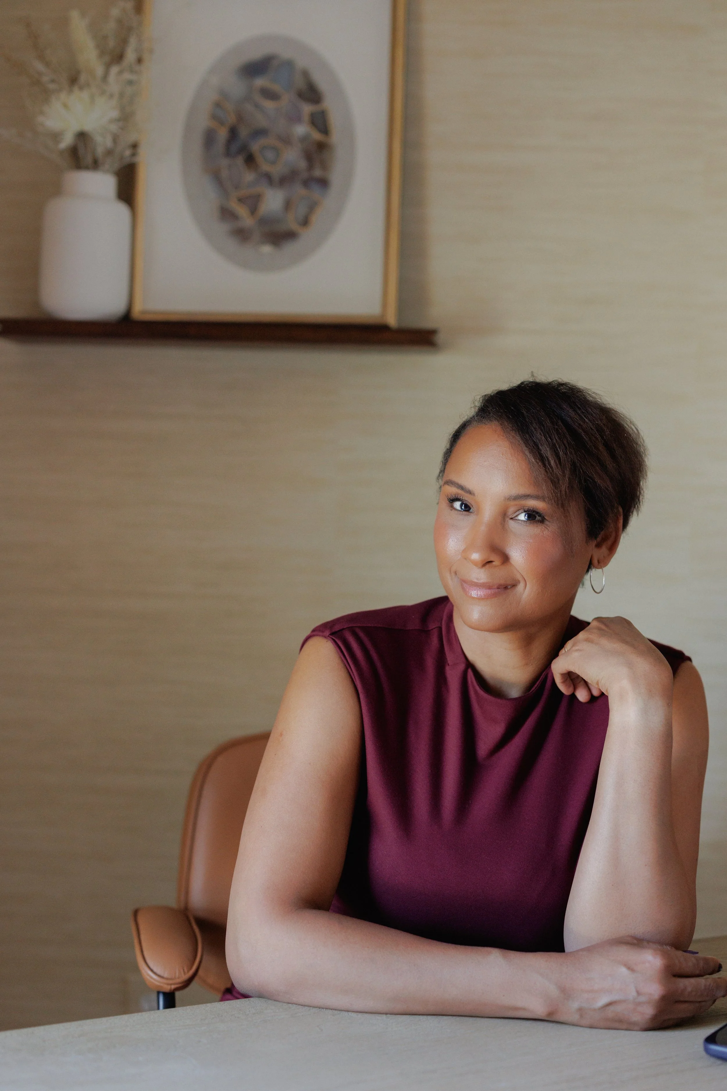 A woman with short brown hair and hoop earrings sits at a table, smiling gently, wearing a maroon sleeveless top. Behind her is a beige wall, a wooden shelf with a white vase of dried flowers, and a framed artwork. Part of a brown chair is visible.