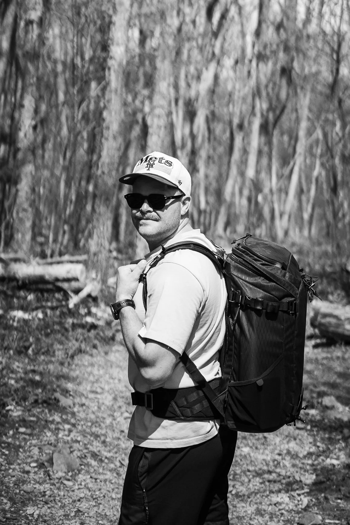 Mann mit Sonnenbrille, Baseballkappe und Rucksack beim Wandern im Wald.
