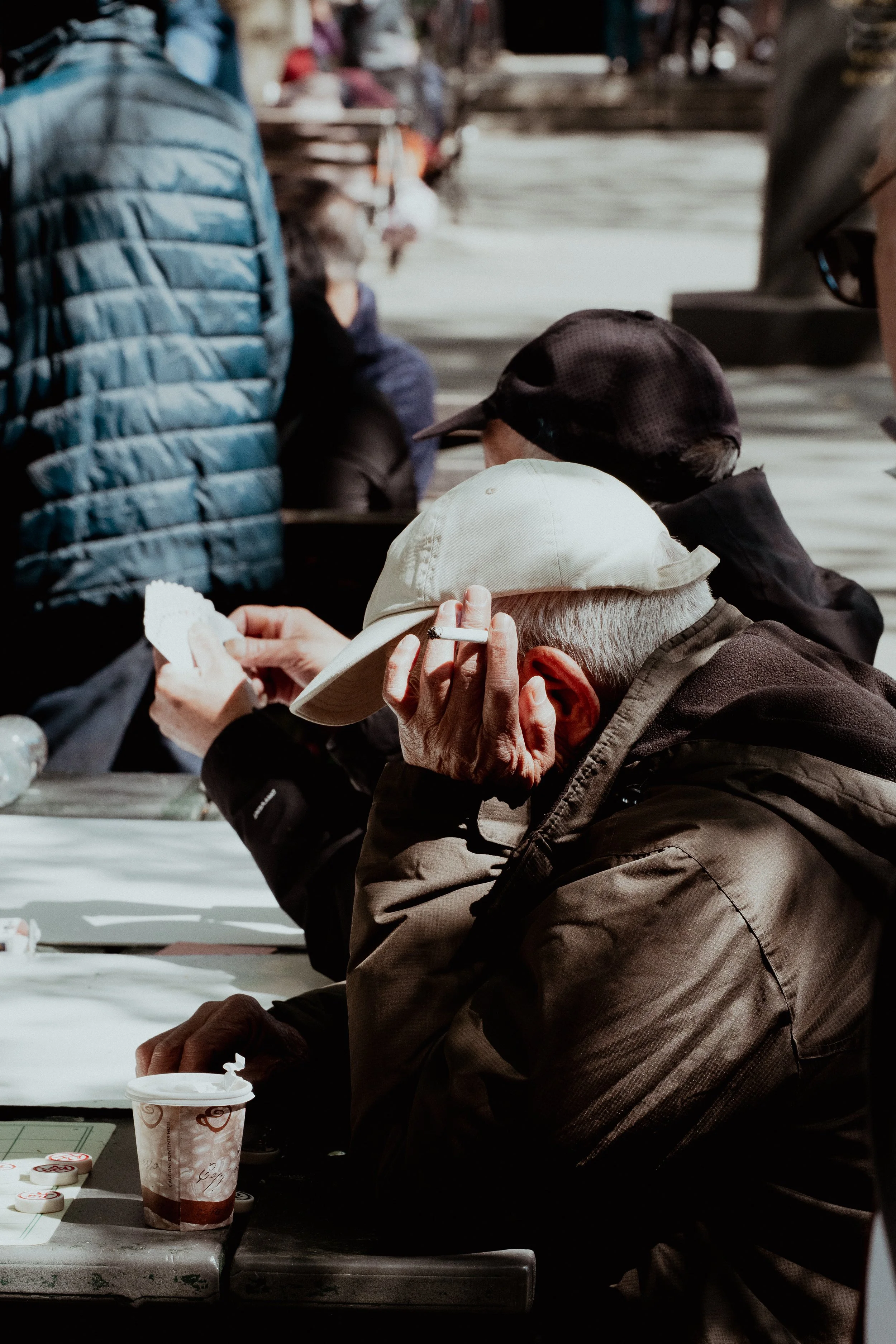 Menschen sitzen an einem Tisch, eine ältere Dame trägt einen Sonnenhut, andere tragen Caps und Jacken, es scheint eine gemeinschaftliche Aktivität zu sein.