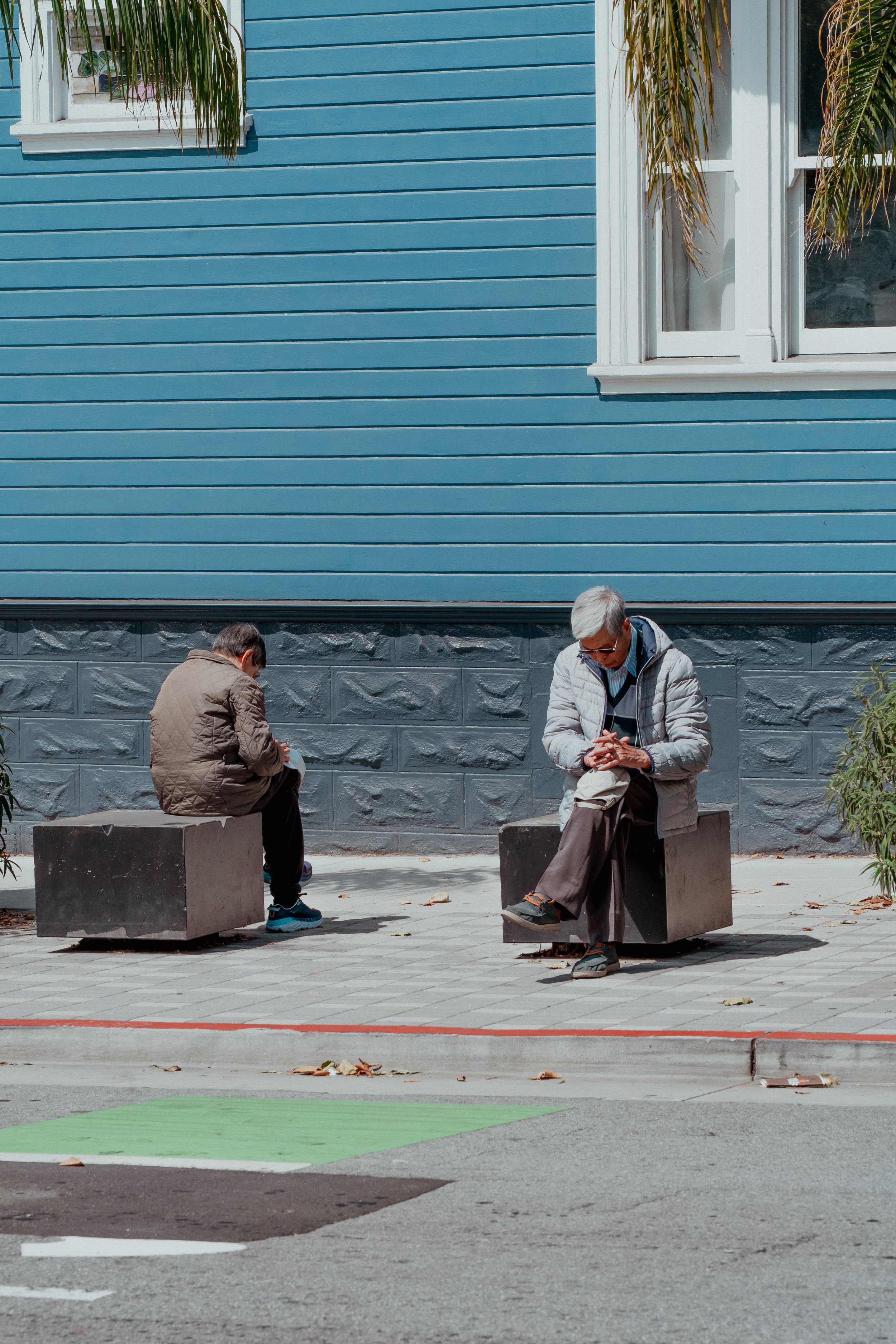 Zwei Männer sitzen auf Betonblöcken vor einem blauen Holzhaus und lesen, während das Wetter sonnig ist.