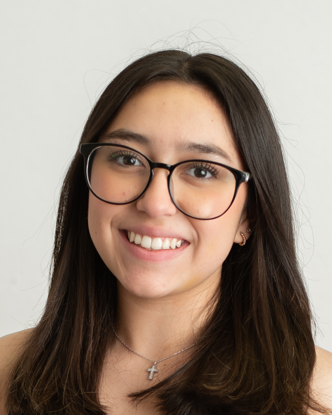 Close-up of a young woman with long brown hair, wearing glasses, smiling, with a cross necklace, and a white background.