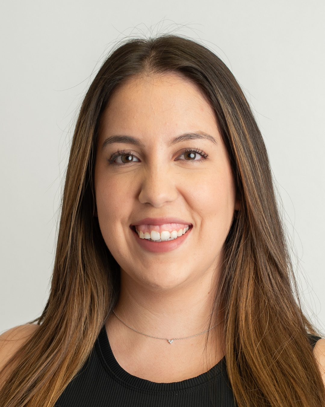 Close-up portrait of a young woman with long brown hair, smiling, wearing a black top and a delicate silver necklace, against a plain light background.